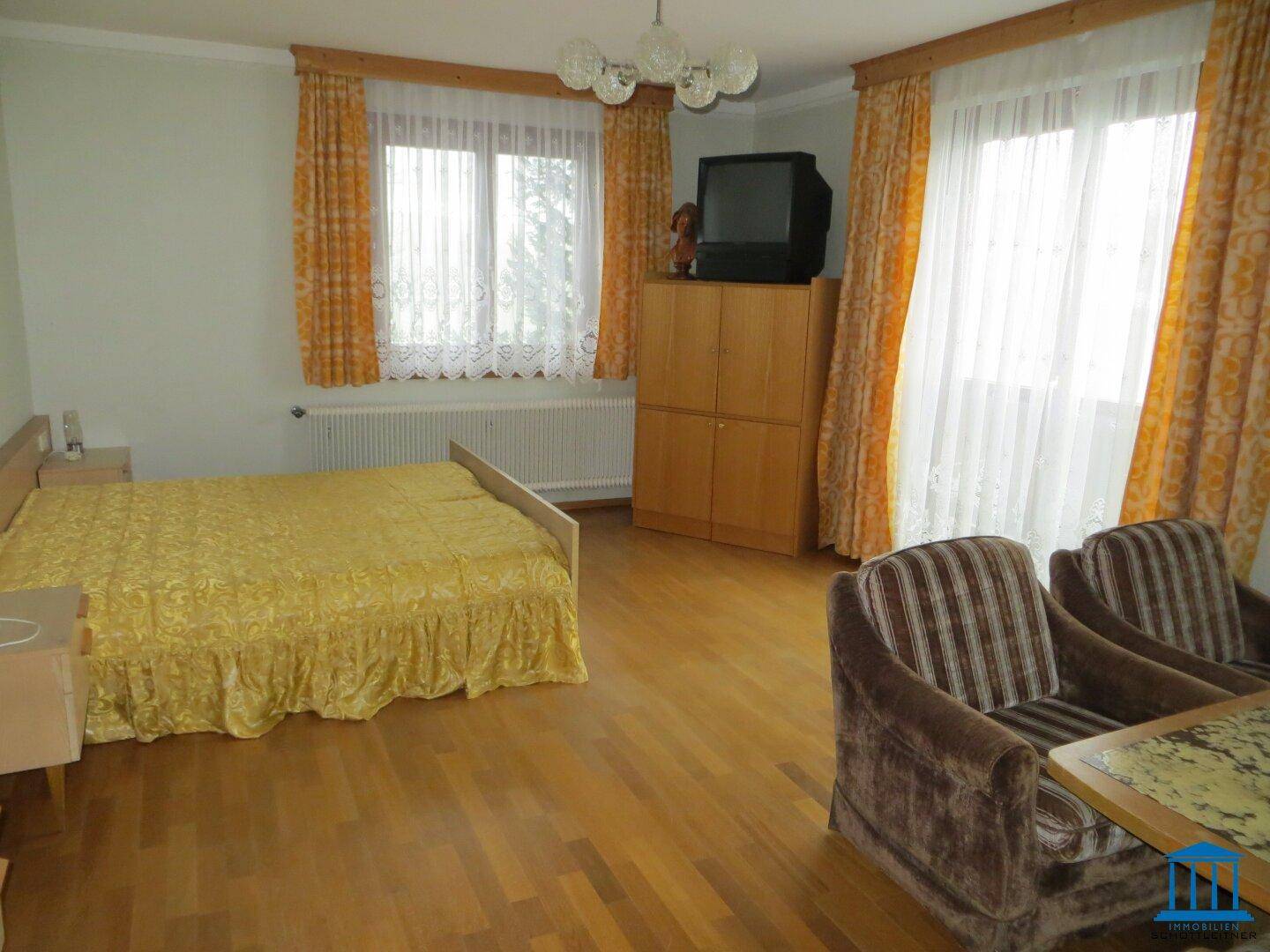 Helles Schlafzimmer mit Doppelbett, Schrank und zwei großen Fenstern mit gelben Vorhängen.
