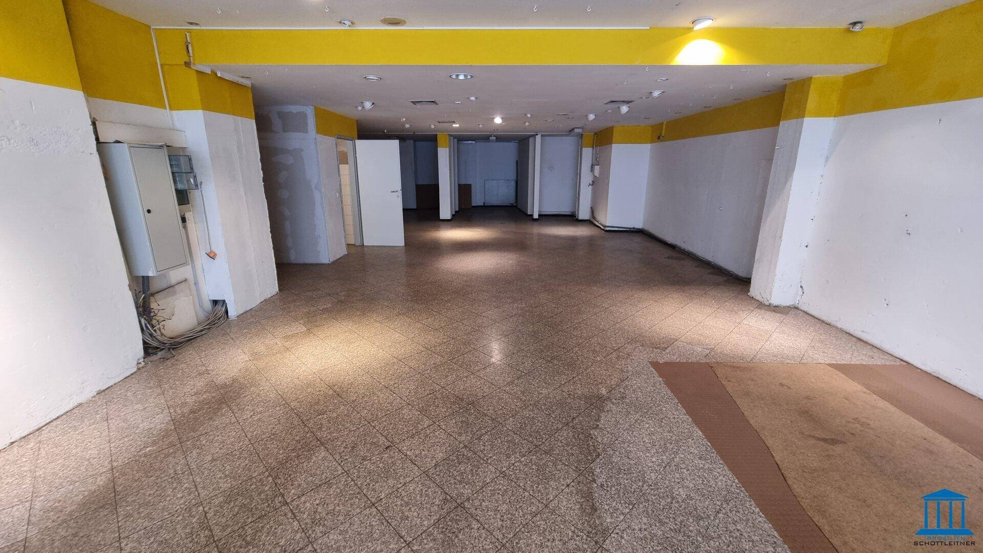 Langer, leerer Ladenraum mit Fliesenboden und gelb-weißen Wänden, der vielseitig nutzbar ist.