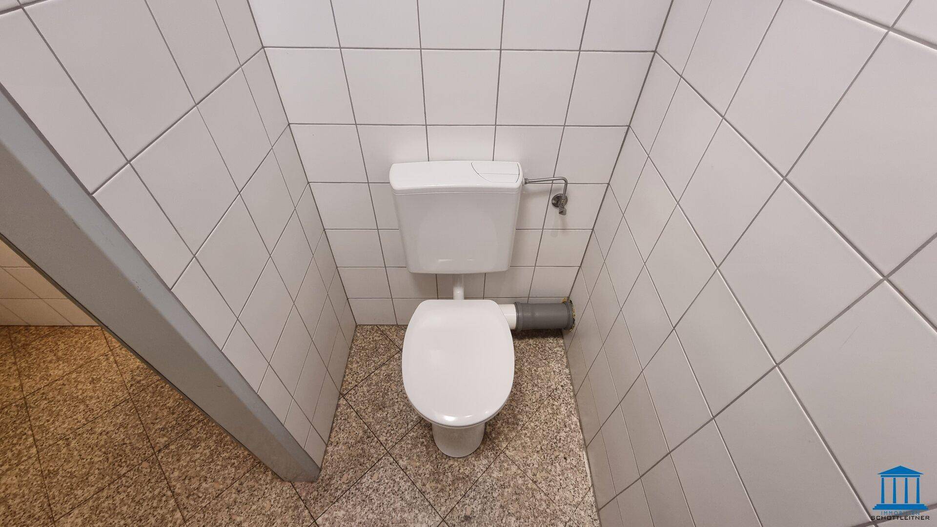 Einfache Toilette mit weißen Wandfliesen und Fliesenboden, in einem funktionalen Zustand.