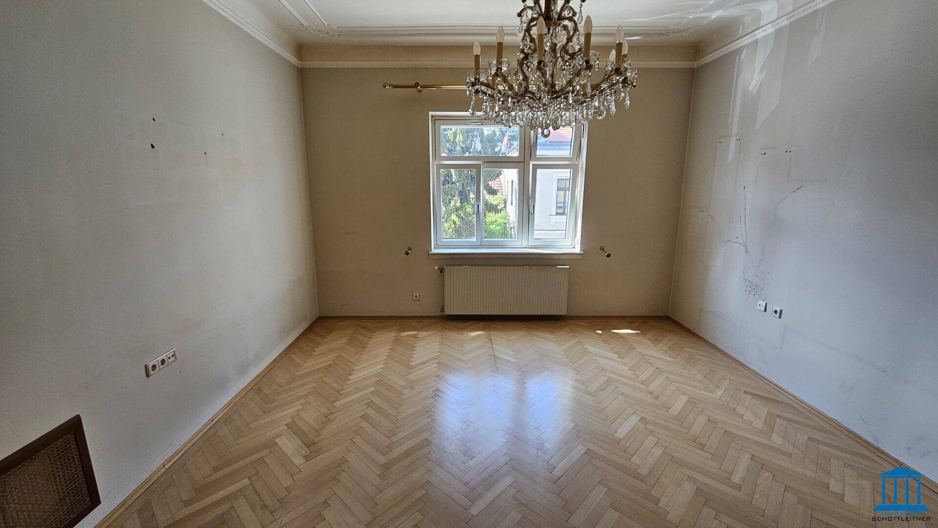 Helles Wohnzimmer mit Fischgrätparkett, einem großen Fenster und einem eleganten Kronleuchter.