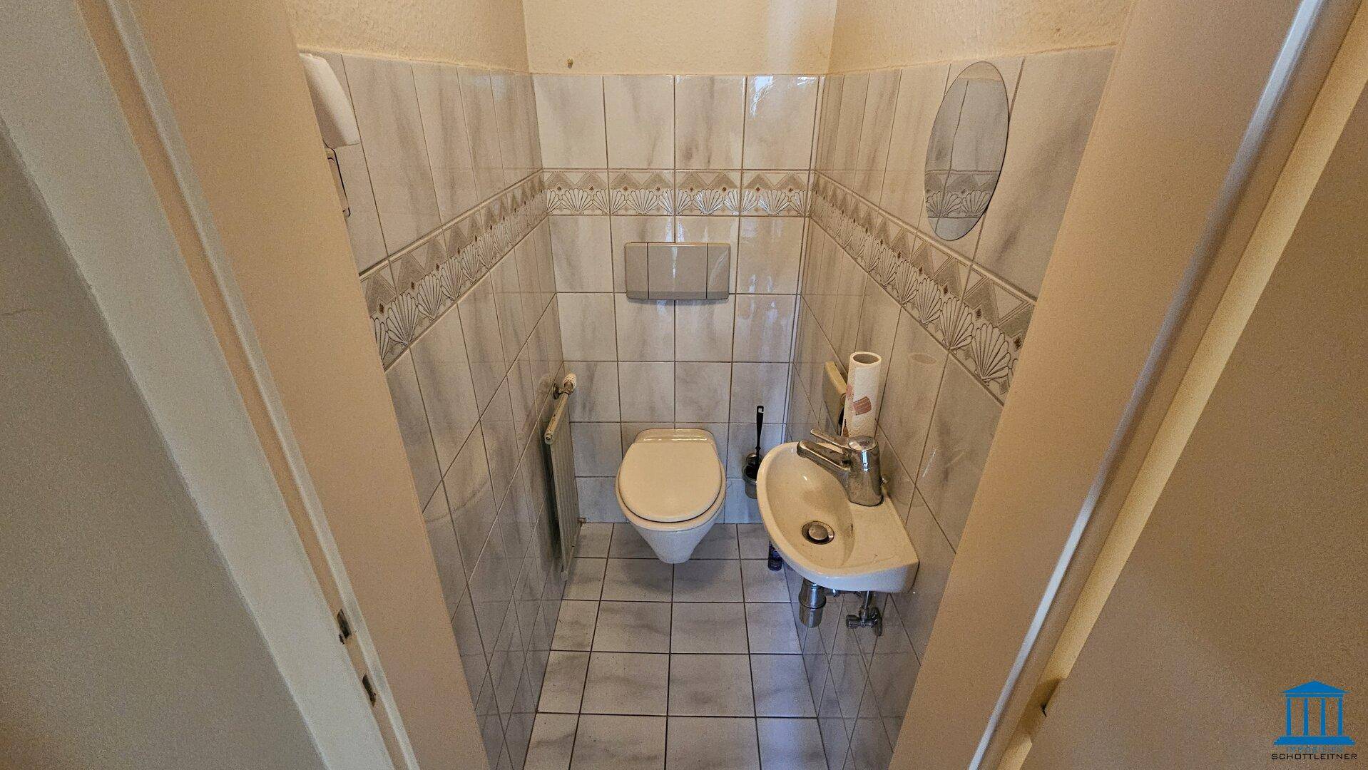 Separate Toilette mit Wand-WC, kleinem Waschbecken und Fliesen an den Wänden.