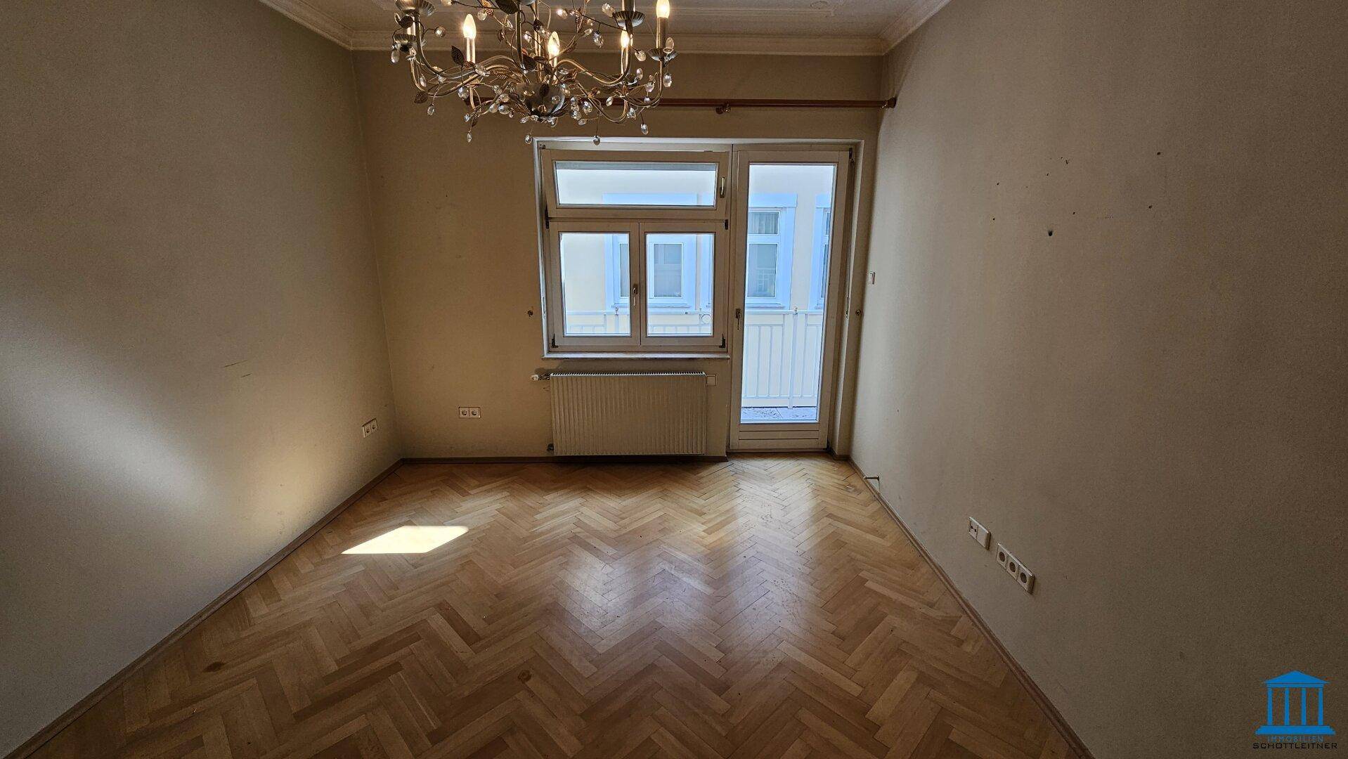Leeres Wohnzimmer mit Fischgrätparkett, Kronleuchter und Zugang zu einem Balkon.