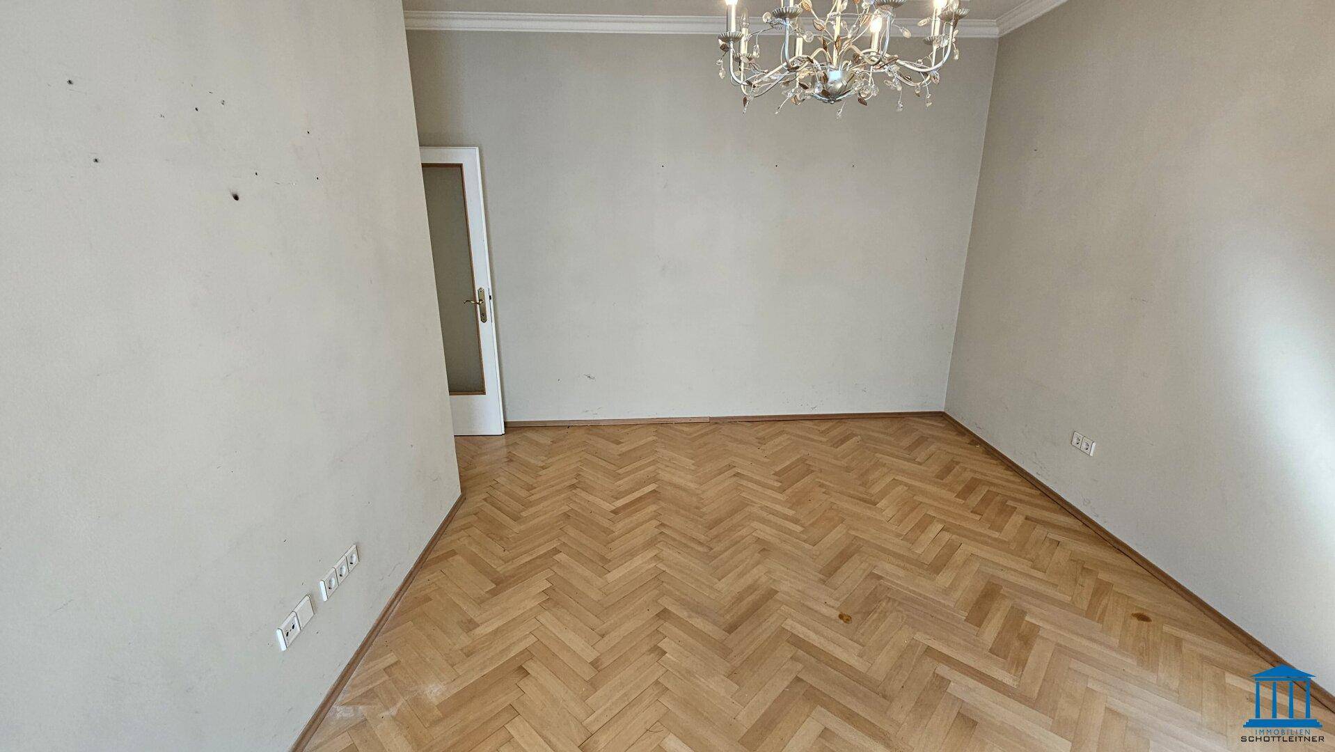 Geräumiges Wohnzimmer mit klassischem Fischgrätparkett und einem eleganten Kronleuchter an der Decke.