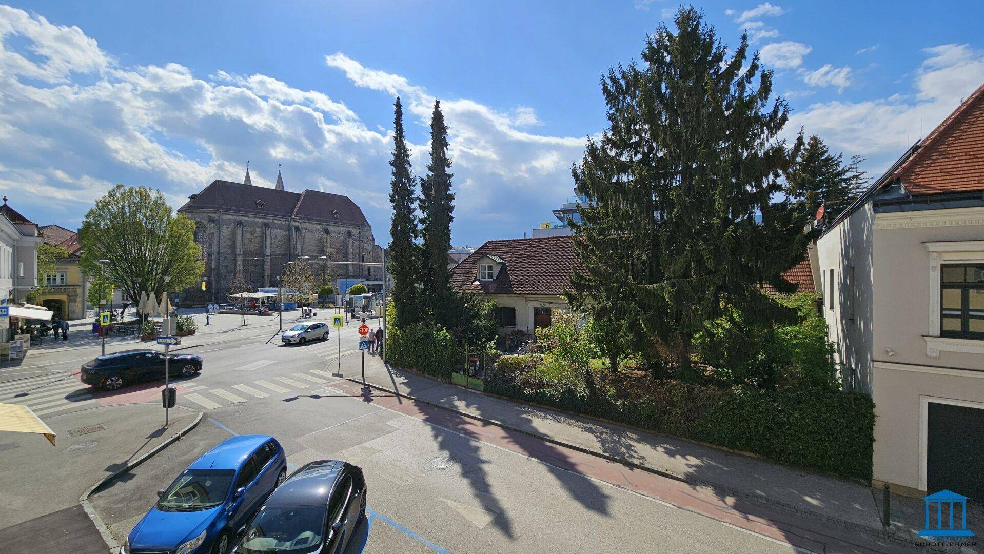 Weitblick über eine Stadtkreuzung mit historischen Gebäuden, Bäumen und geparkten Autos unter blauem Himmel.