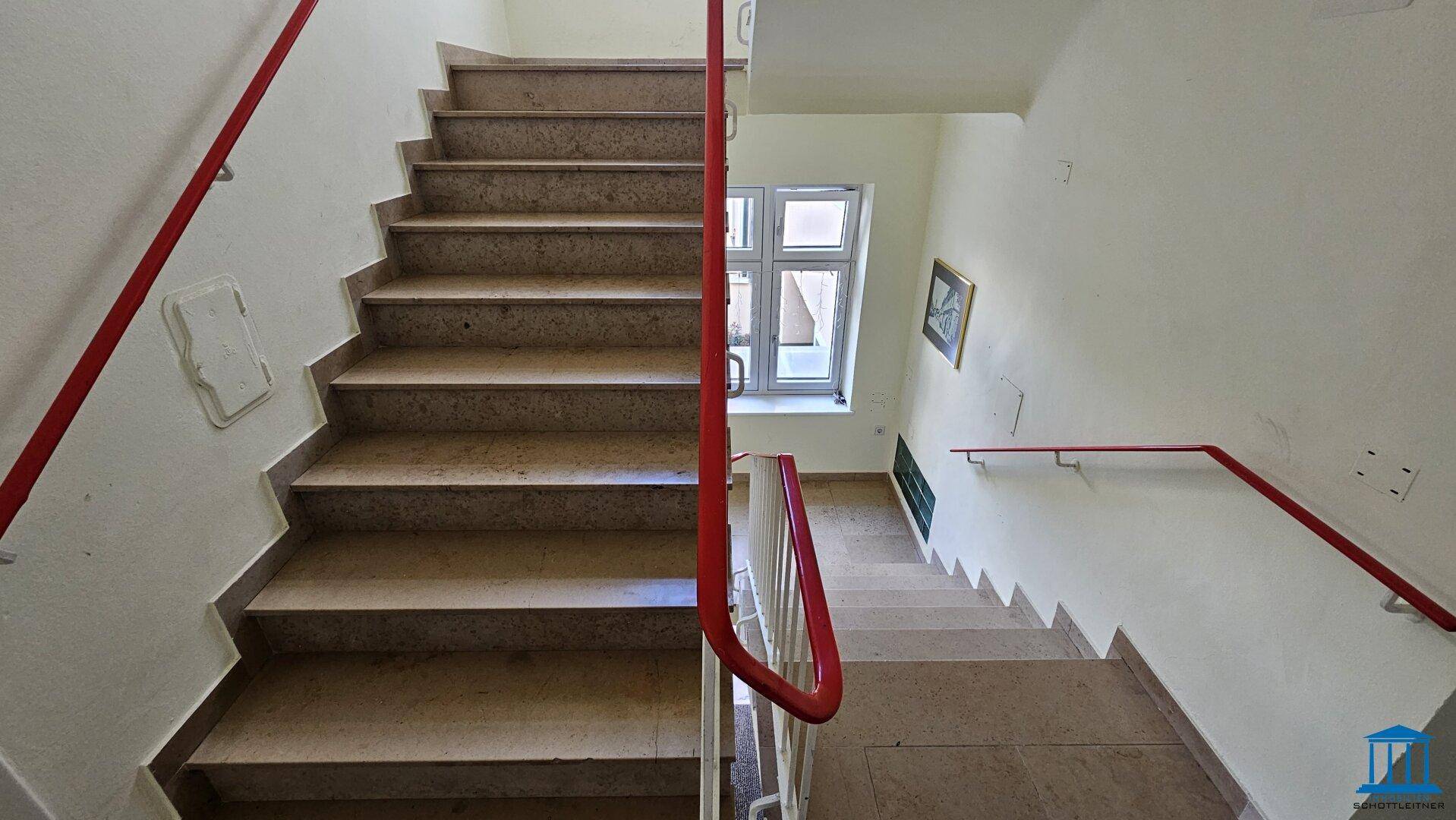 Breiter Treppenaufgang mit Steinstufen und rotem Handlauf, der viel natürliches Licht erhält.
