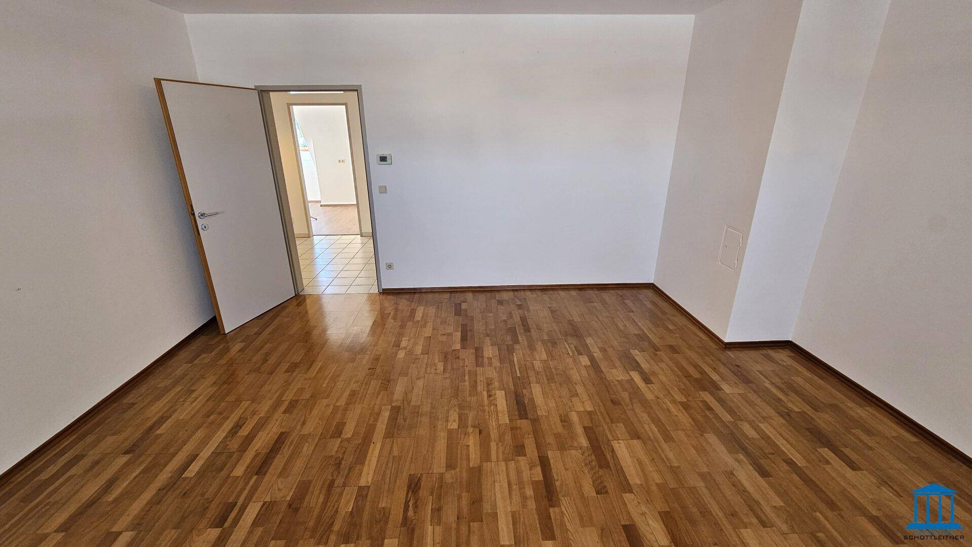 Geräumiges Zimmer mit Holzboden und weiß gestrichenen Wänden, Zugang zu einem gefliesten Bereich.
