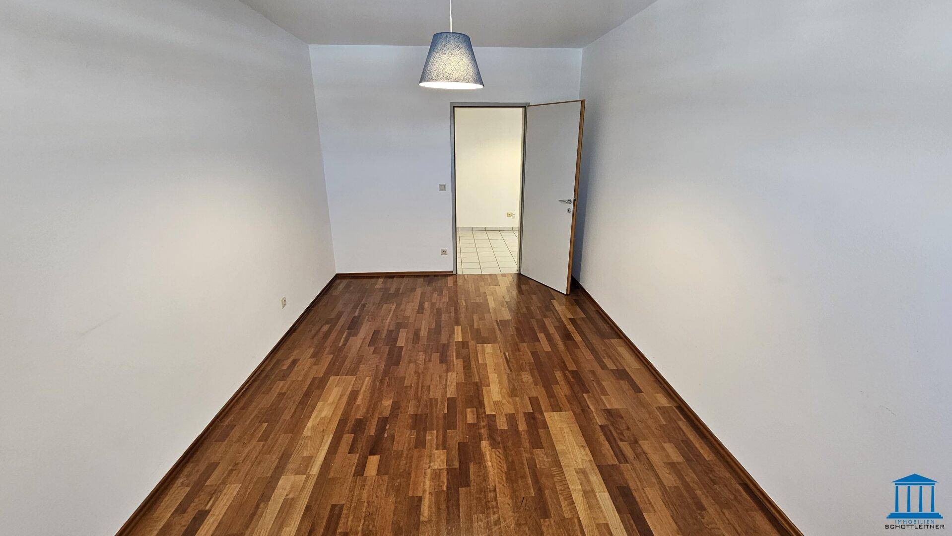 Helles Zimmer mit Parkettboden und weiß gestrichenen Wänden, bietet Zugang zu einem gefliesten Bereich.