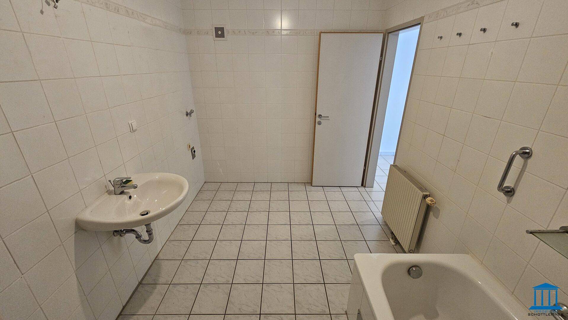 Funktionales Badezimmer mit weißen Fliesen, Waschbecken und Heizkörper, Zugang zu weiteren Räumen.