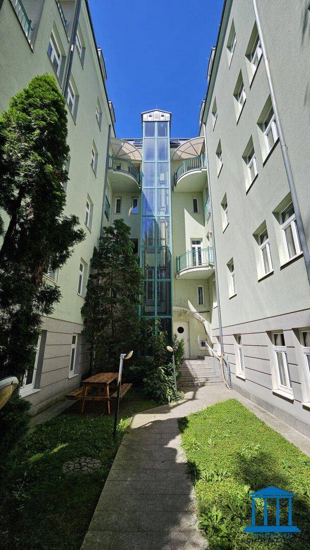 Attraktive Gebäudefassade mit zentralem Glasaufzug und Balkonen, umgeben von viel Grün.