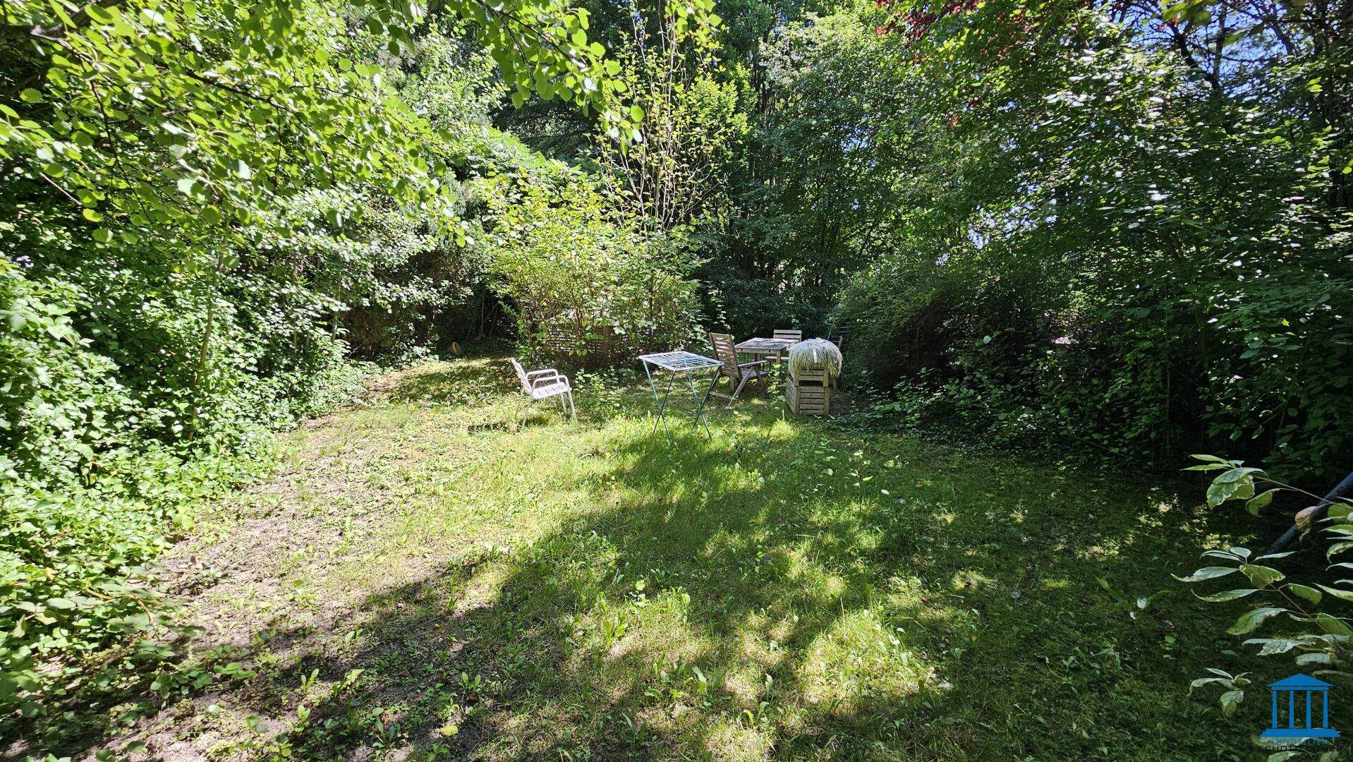 Naturbelassener Gartenbereich mit Sitzgelegenheiten, umgeben von üppigem Grün und Bäumen.