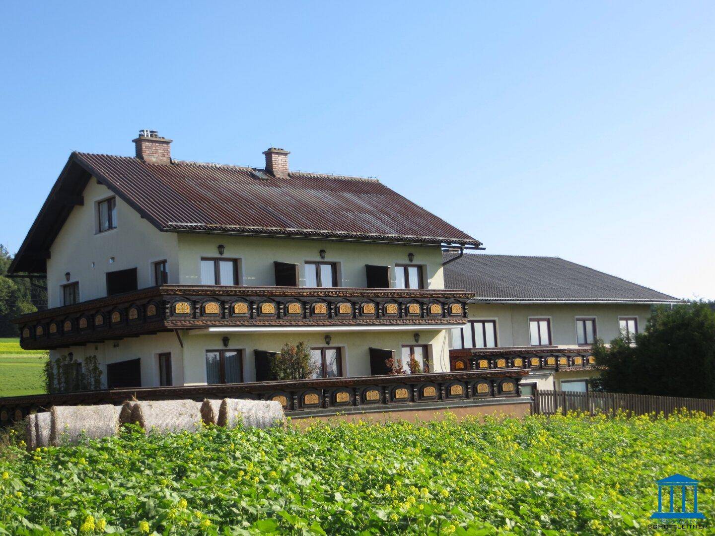 Ansicht des traditionellen Gebäudes mit Holzbalkonen, eingebettet in eine ländliche Landschaft mit weiten Feldern.