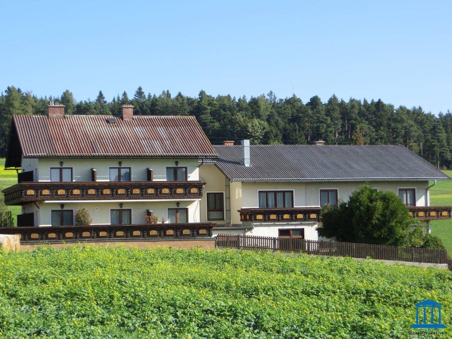 Traditionelles Gebäude mit mehreren Balkonen, umgeben von grünen Feldern und Wäldern unter blauem Himmel.