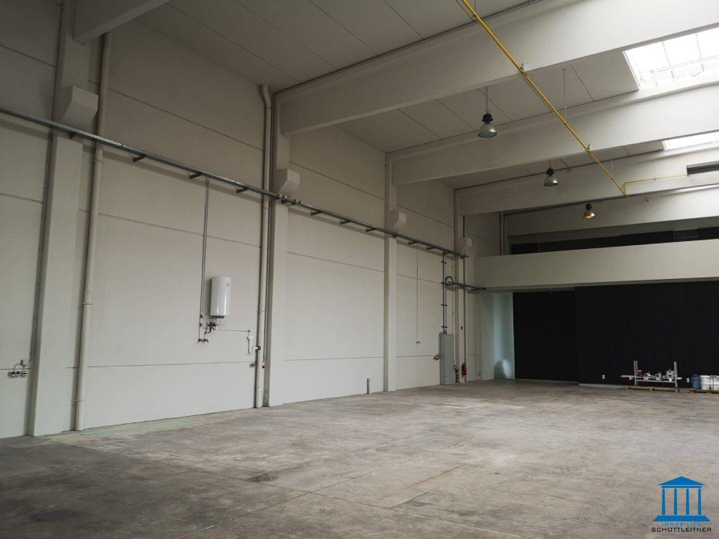 Leere Lagerhalle mit hohen Wänden, Betonboden und sichtbaren Installationen an der Wand.