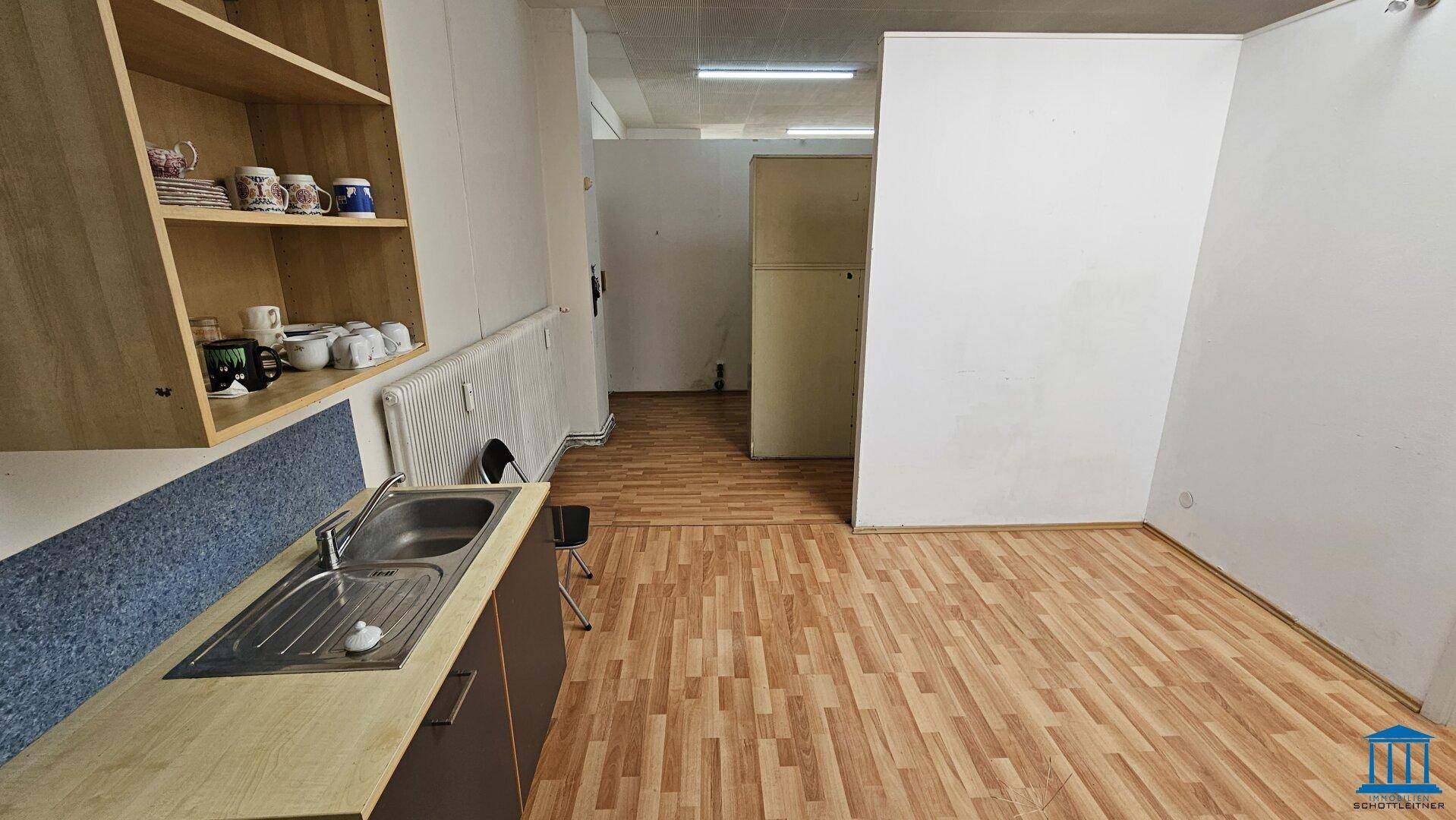 Küchenzeile mit Spülbecken und Regalen, die in einen größeren Raum mit Laminatboden übergeht.