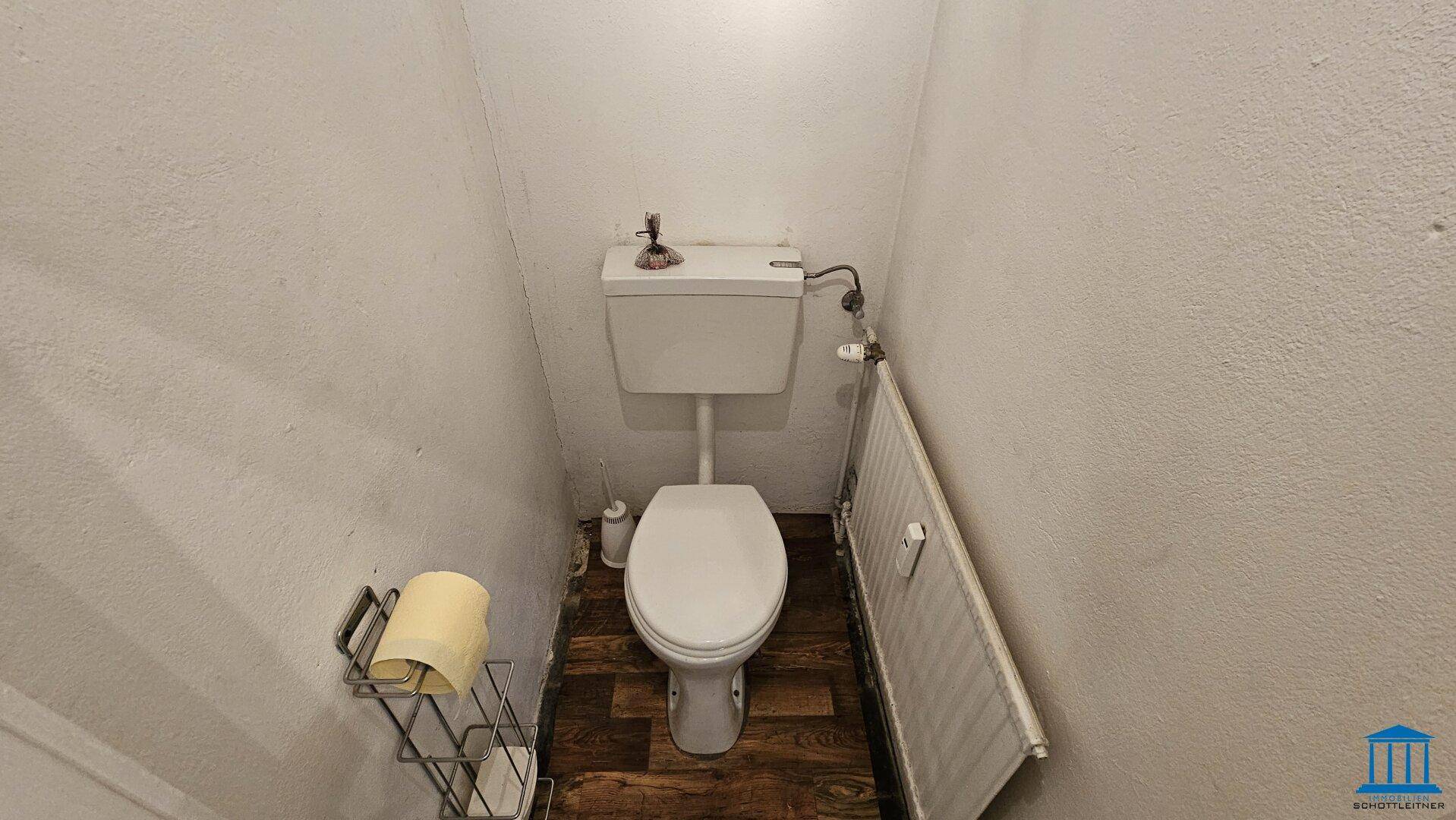 Separate Toilette mit WC, Heizkörper und dunklem Bodenbelag in einem schlichten Raum.
