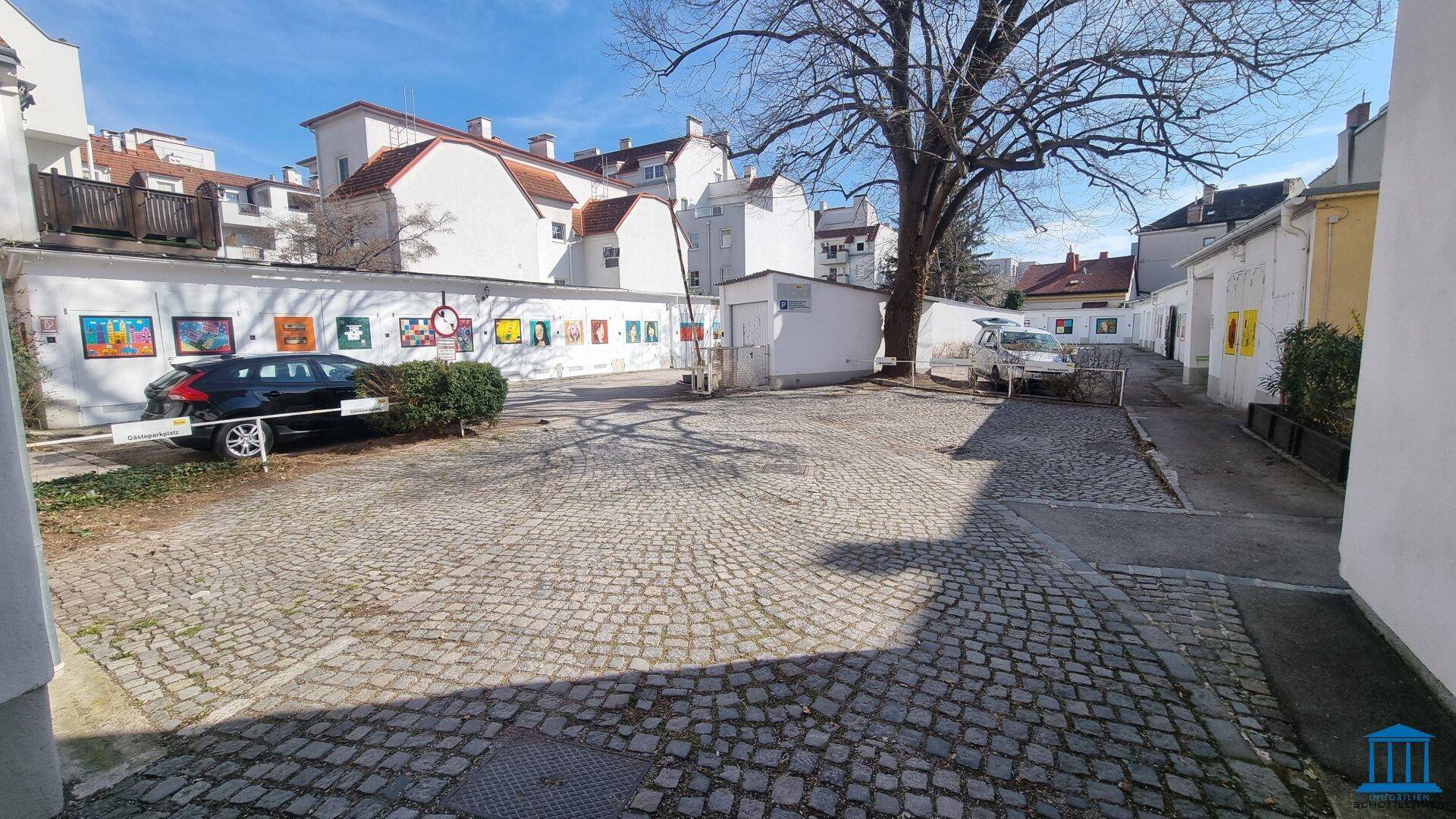 Gepflasterter Innenhof mit parkenden Autos und einem großen Baum, umgeben von Gebäuden.