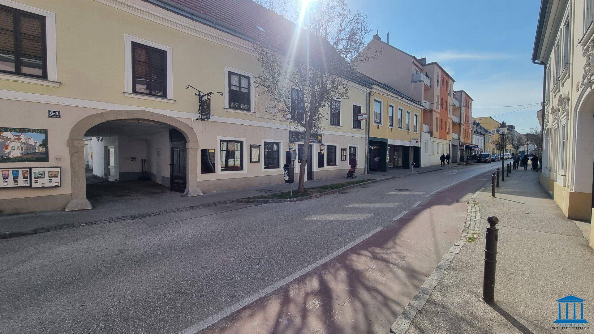 Belebte Straßenansicht mit einer Reihe von Gebäuden, geparkten Fahrzeugen und einem sonnigen Himmel.