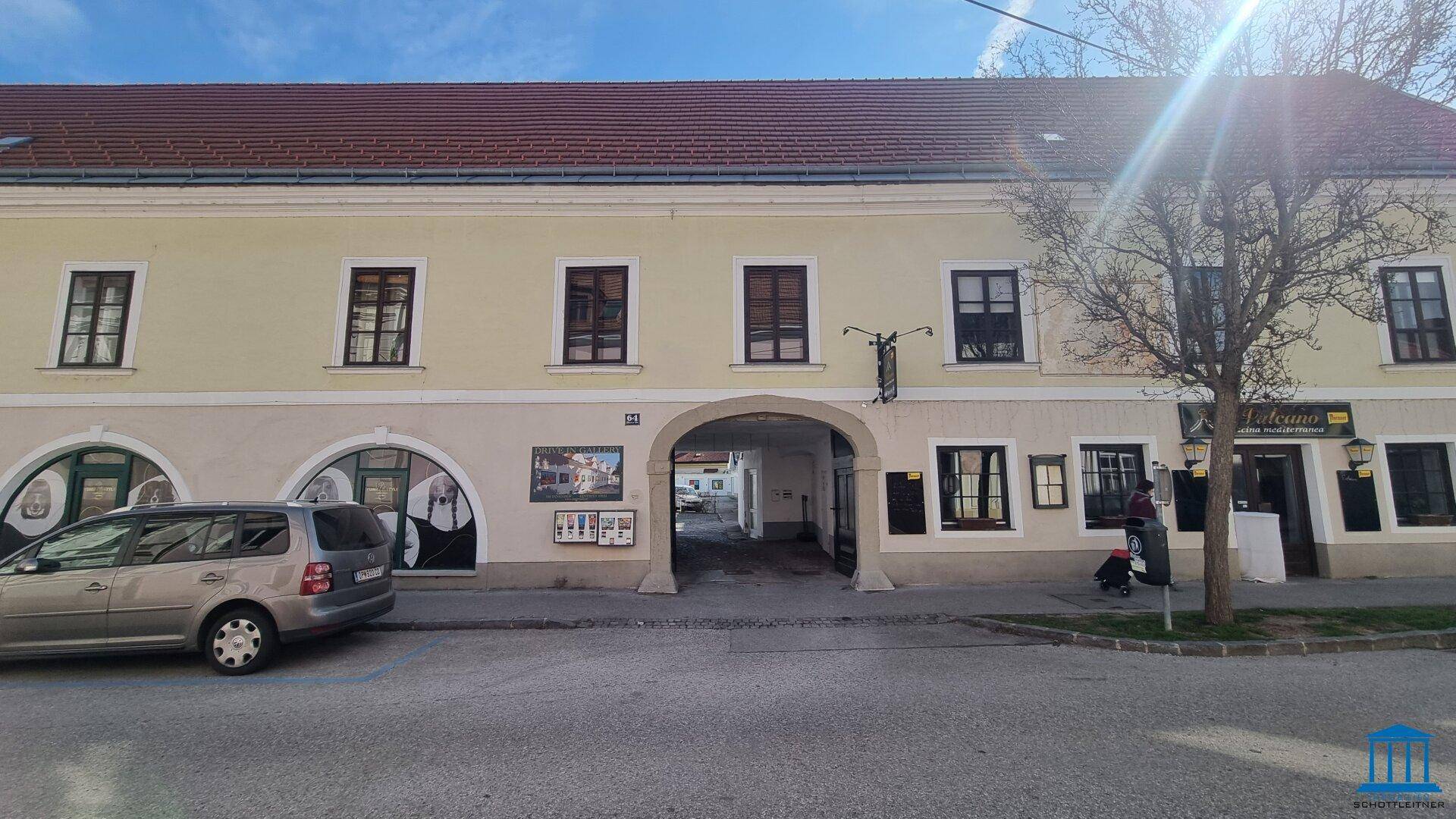 Straßenansicht eines Gebäudes mit einem Torbogen, der zu einem Innenhof führt, und einem geparkten Auto.