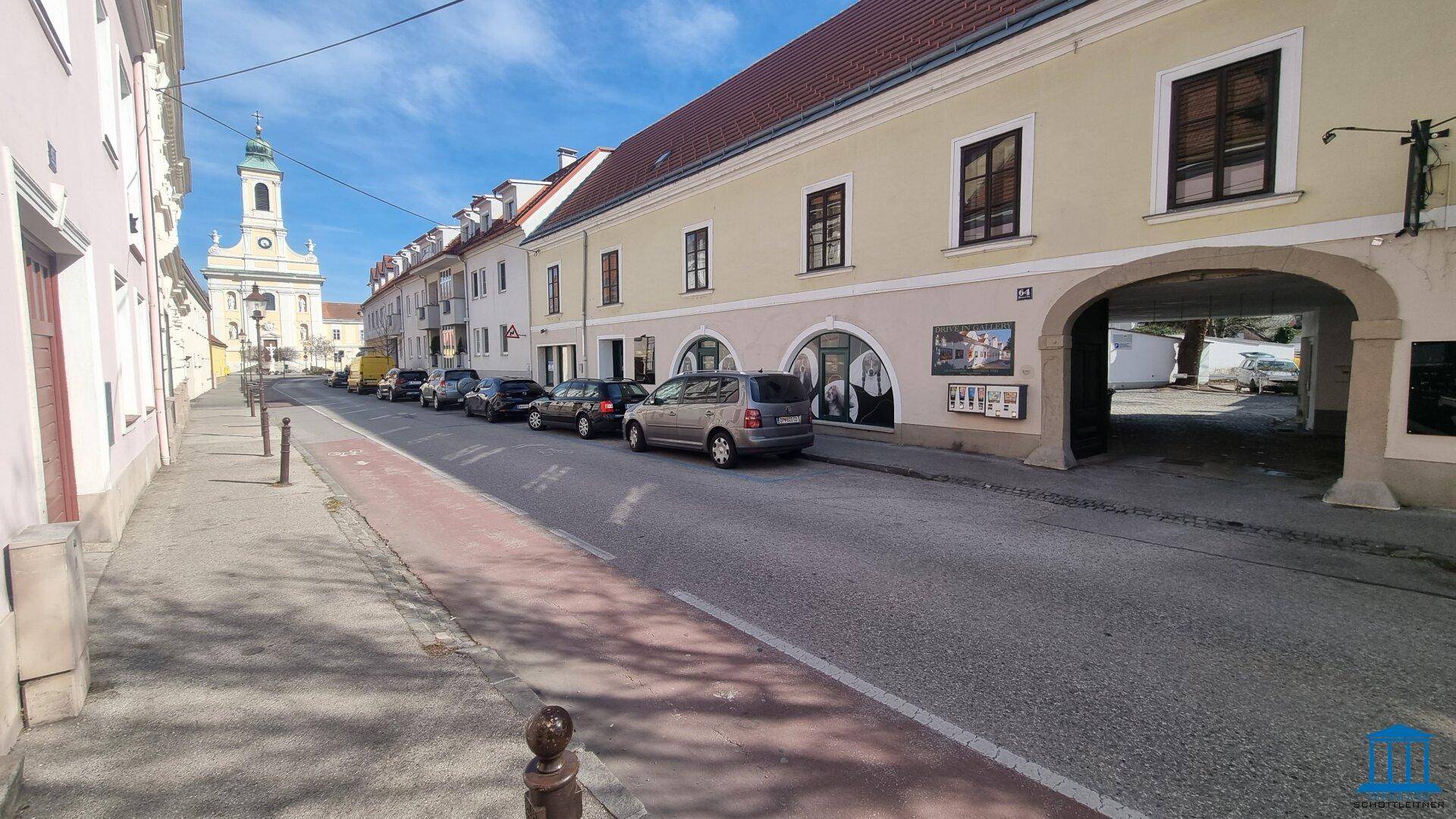 Weitläufige Straßenansicht mit historischen Gebäuden, geparkten Autos und einem Kirchturm in der Ferne.