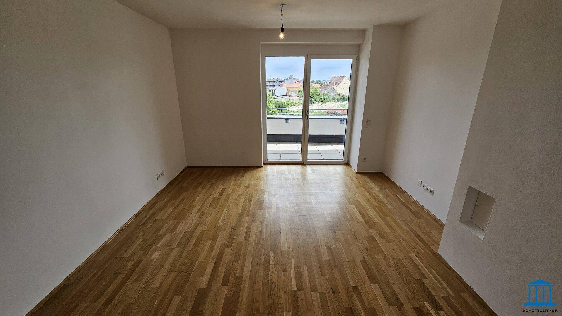 Helles, leeres Wohnzimmer mit Parkettboden und Zugang zu einem Balkon.
