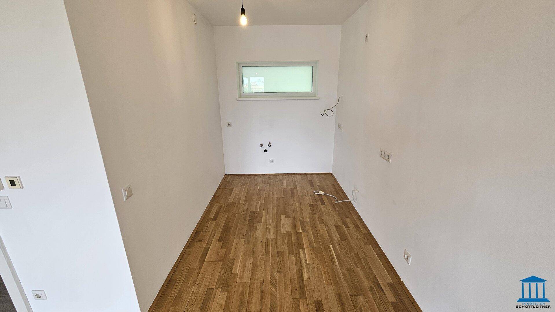 Leerer Küchenraum mit Parkettboden, Fenster und Anschlüssen für eine zukünftige Einbauküche.