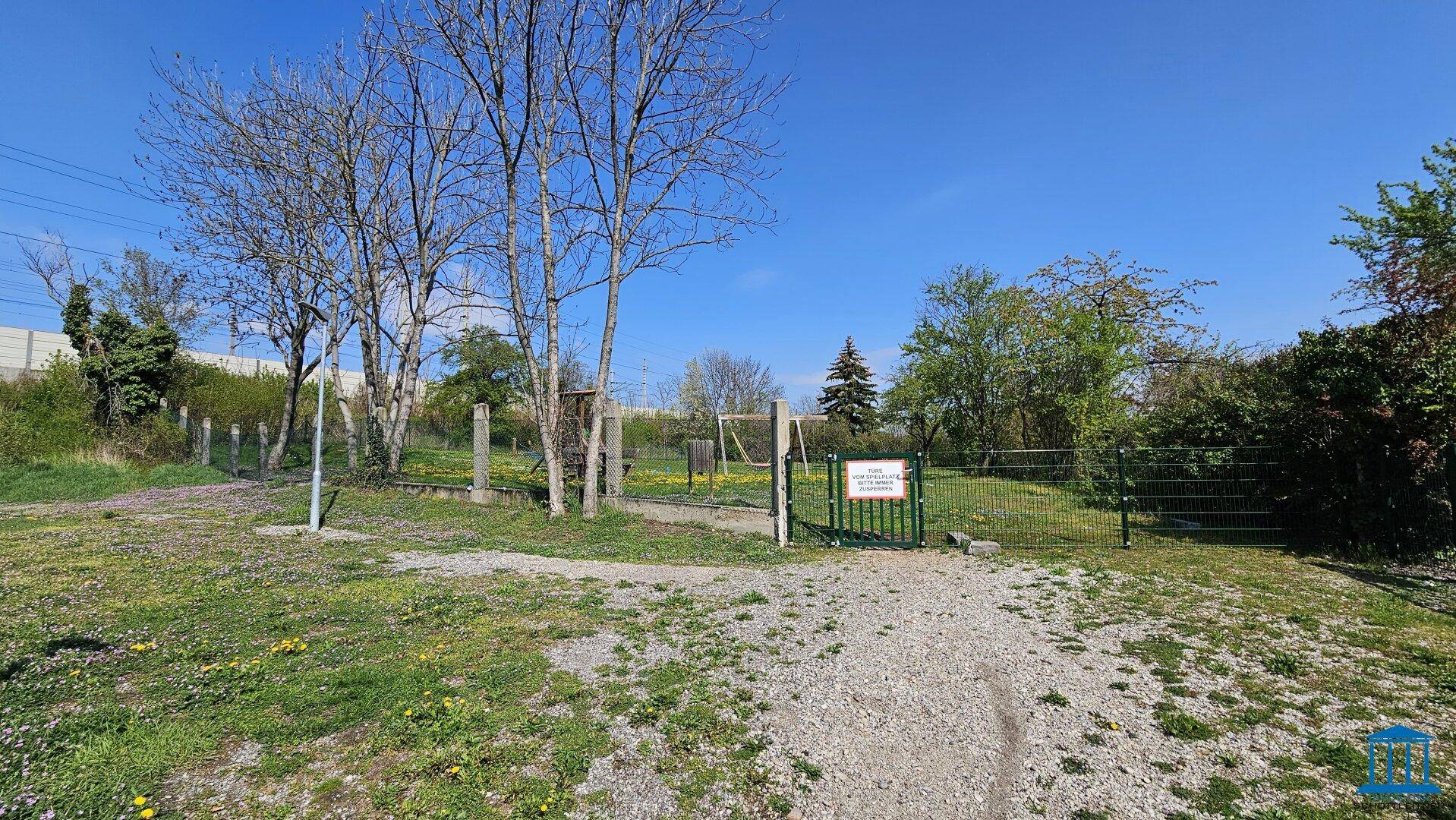 Eingang zu einem umzäunten Spielplatz mit Bäumen und Grünflächen, ideal für Kinder.