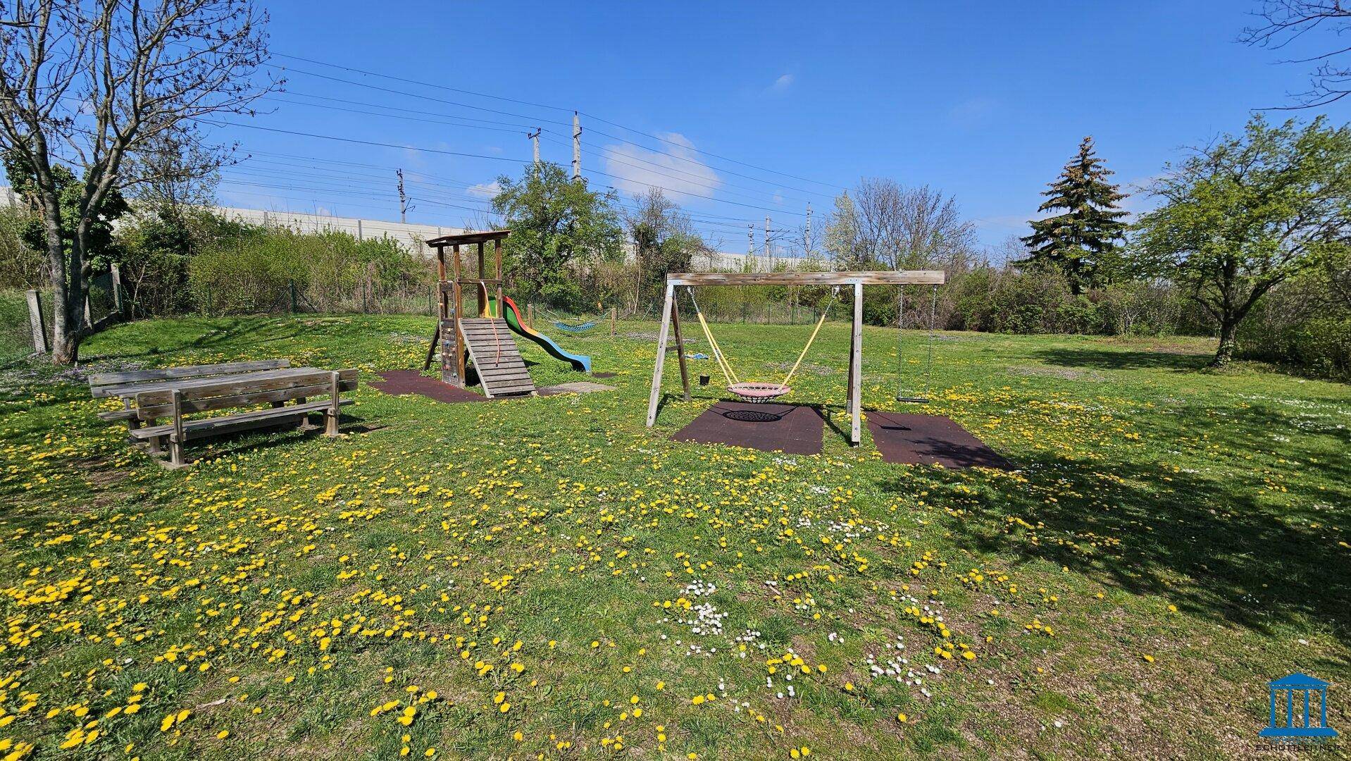 Kinderspielplatz mit Rutsche, Schaukel und Sitzbank, umgeben von einer grünen Wiese.