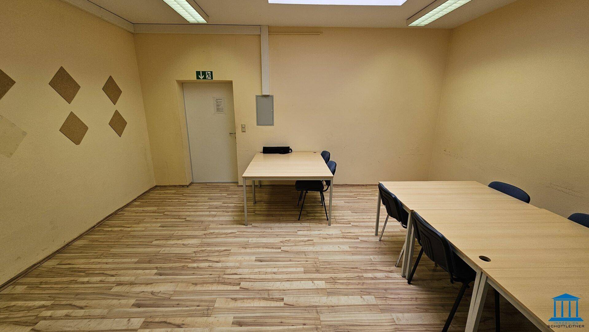 Einfacher Büroraum mit Holzboden, zwei Tischen und Stühlen, sowie einer Tür mit Notausgangsschild.