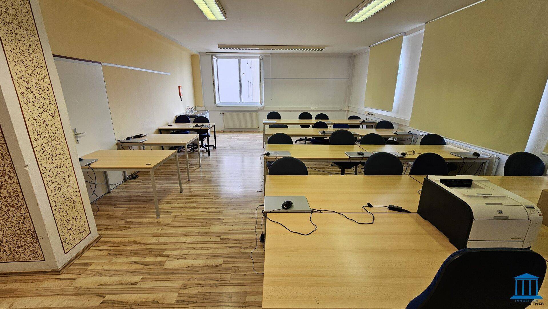 Heller Büroraum mit Fenster, ausgestattet mit mehreren Arbeitsplätzen und einem Drucker, ideal für produktives Arbeiten.