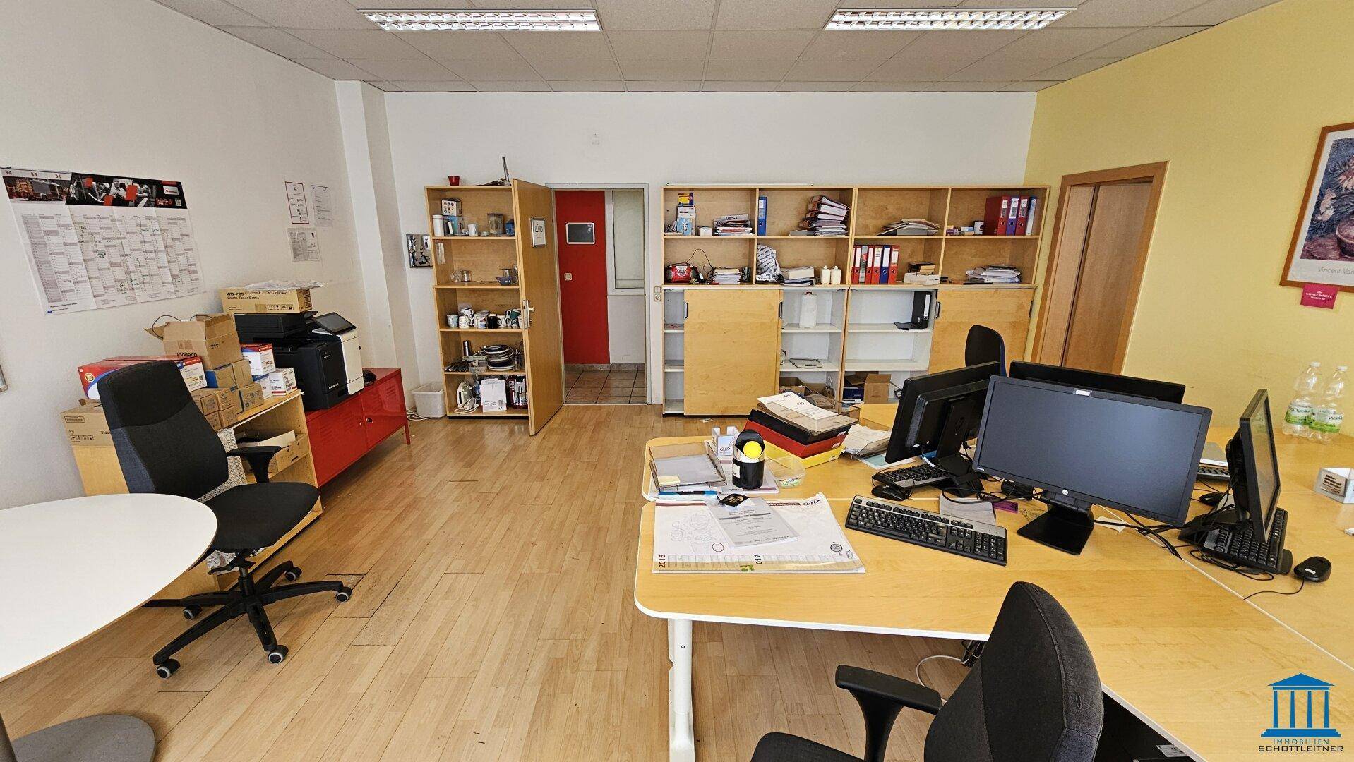 Großraumbüro mit mehreren Arbeitsplätzen, Computern und Regalen für Akten und Büromaterial.