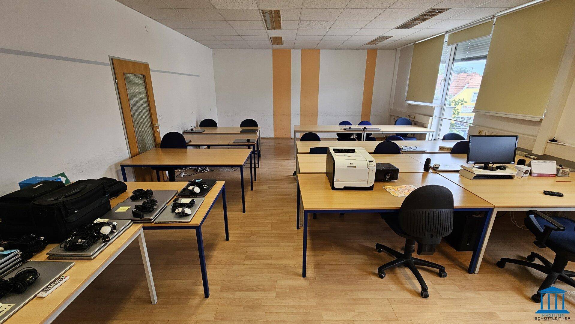 Großer Büroraum mit mehreren Arbeitsplätzen, Computern und einem Drucker, gut beleuchtet.