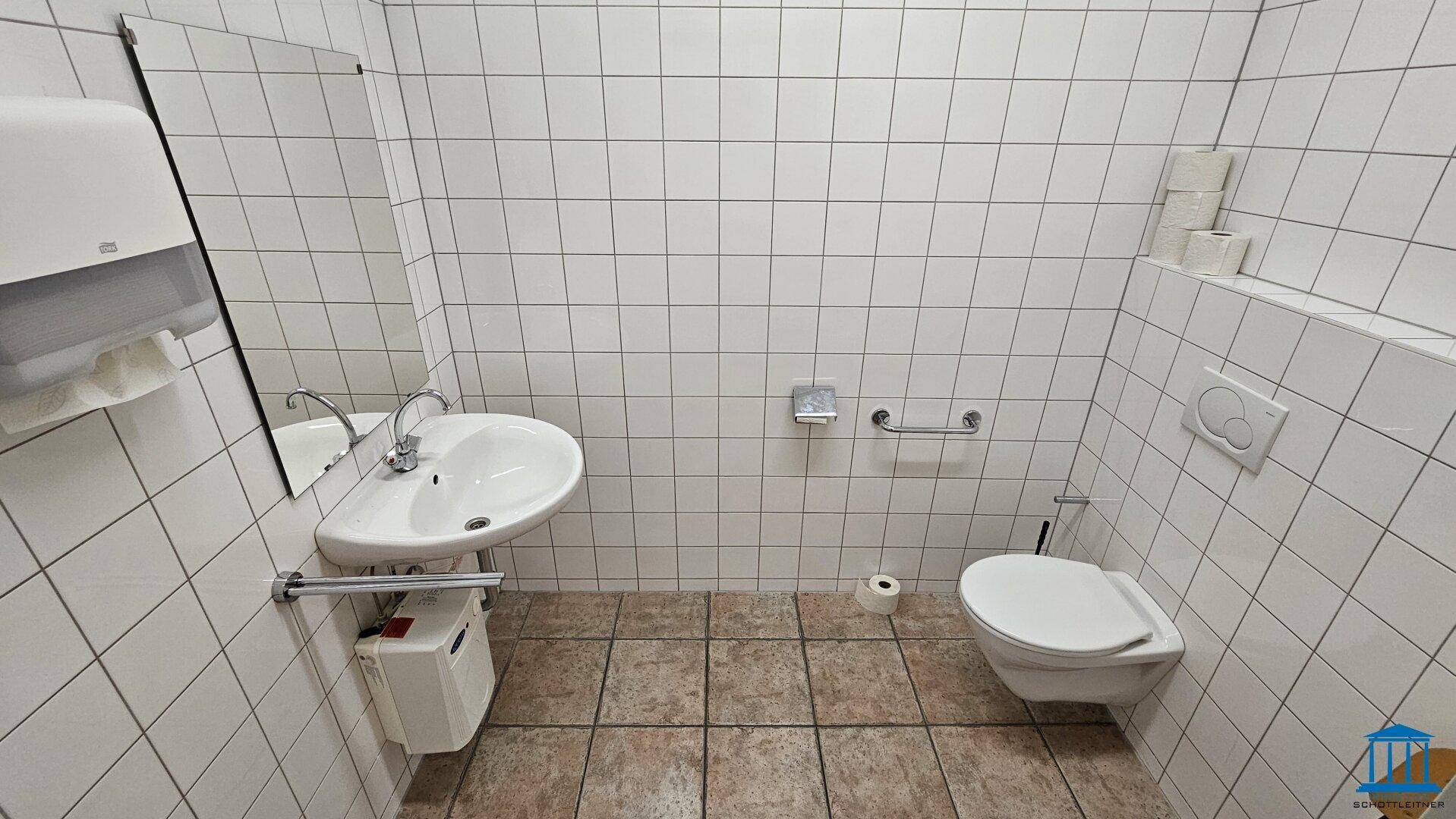 Badezimmer mit weißen Fliesen, Waschbecken, Toilette und Haltegriffen für Barrierefreiheit.