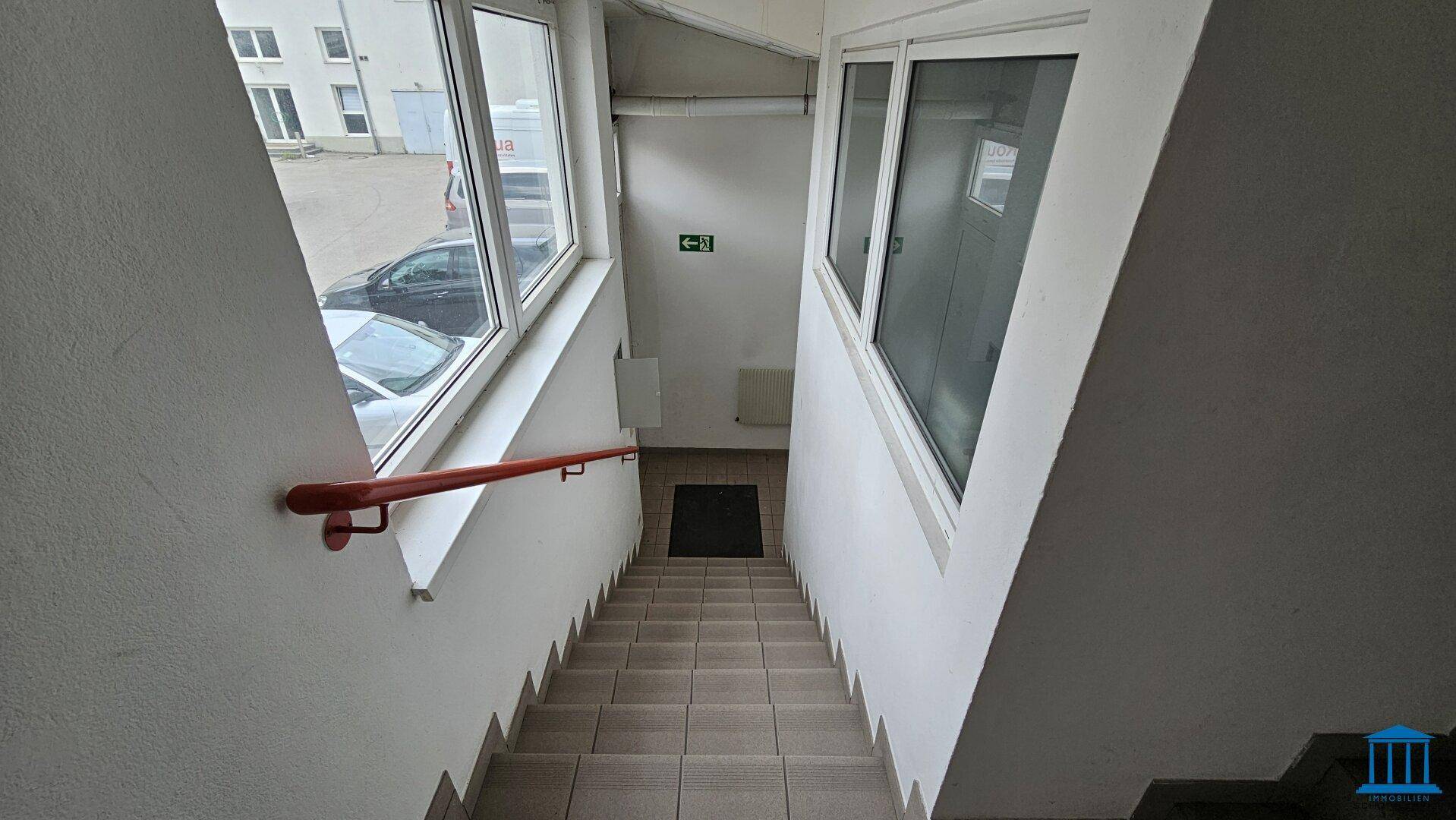 Treppenabgang mit Fliesenstufen, rotem Handlauf und großen Fenstern, die viel Licht hereinlassen.