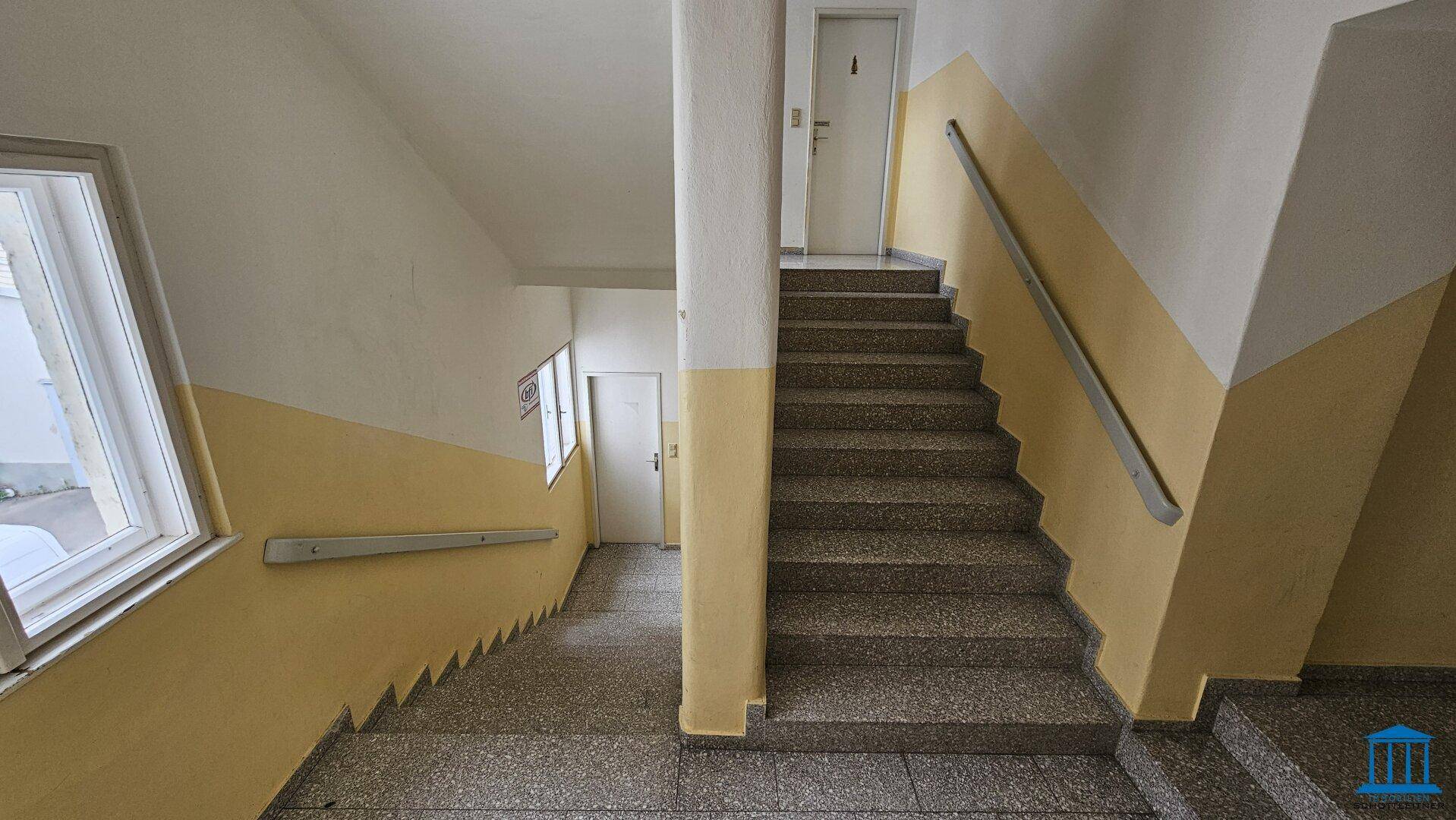 Treppenhaus mit robusten Steinstufen und Handlauf, Wände in Gelb und Weiß gestrichen, mit Fenster für Tageslicht.