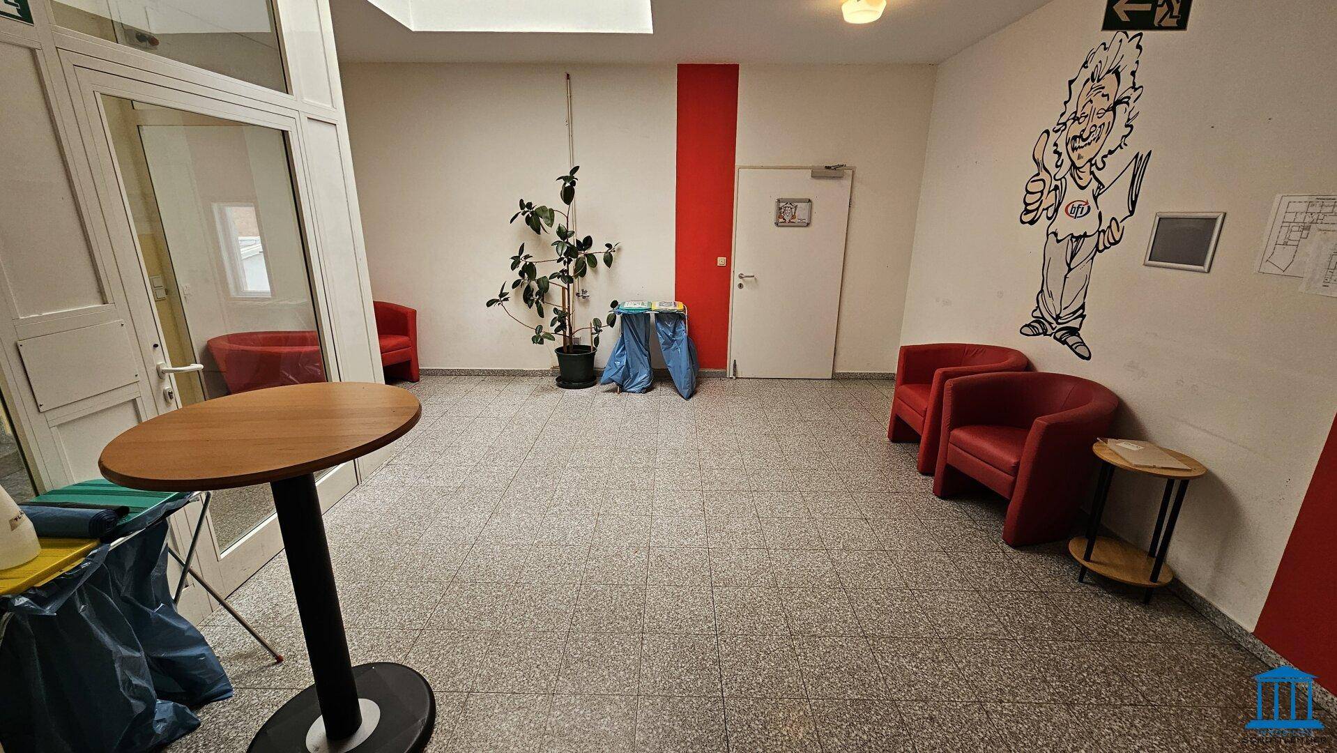 Geräumiger Flur mit Fliesenboden, roten Sesseln und einem Stehtisch, dekoriert mit einer Pflanze und Wandkunst.