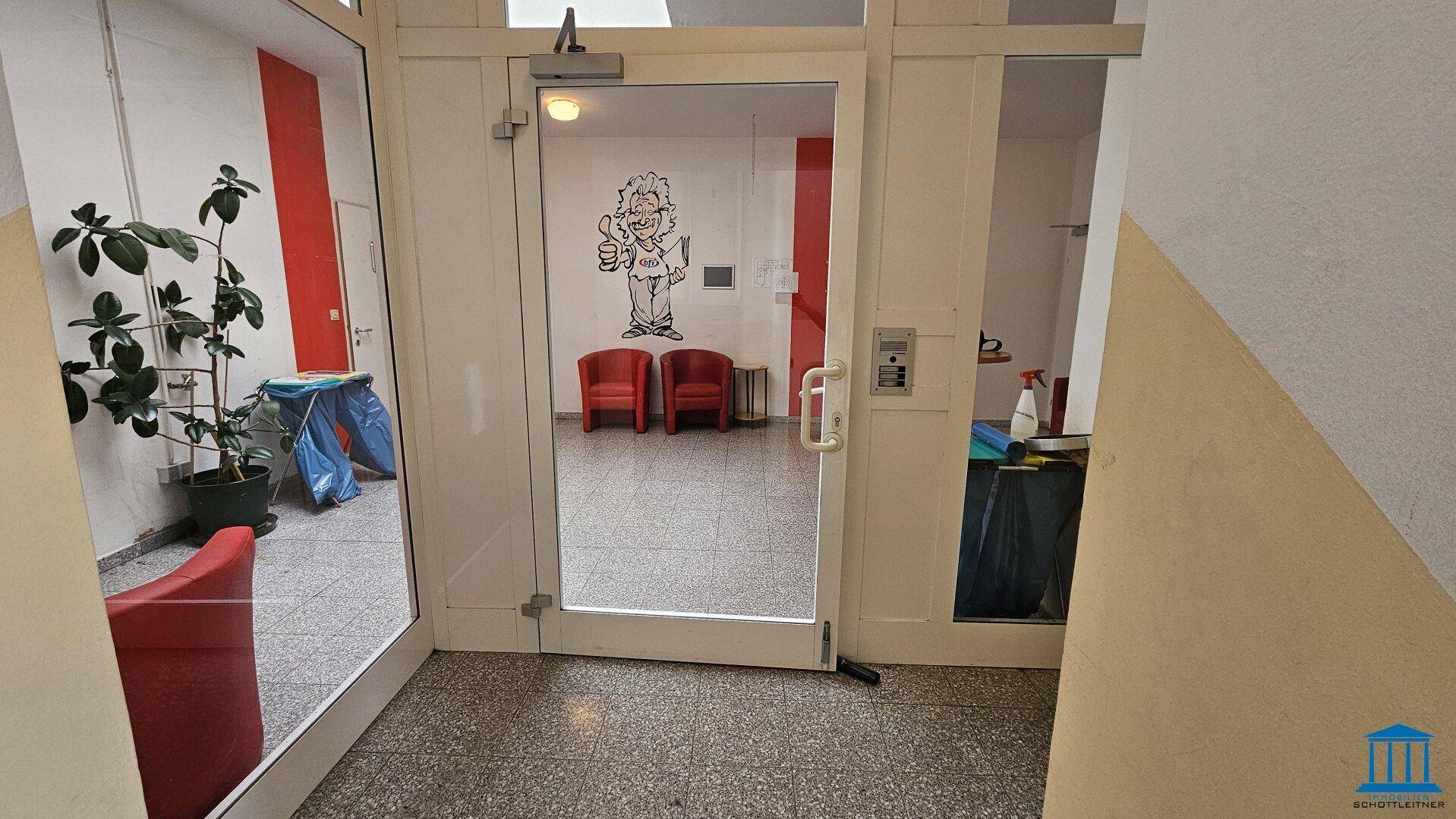 Eingangsbereich mit Glastür und Sitzgelegenheiten, bietet Zugang zum Gebäude.