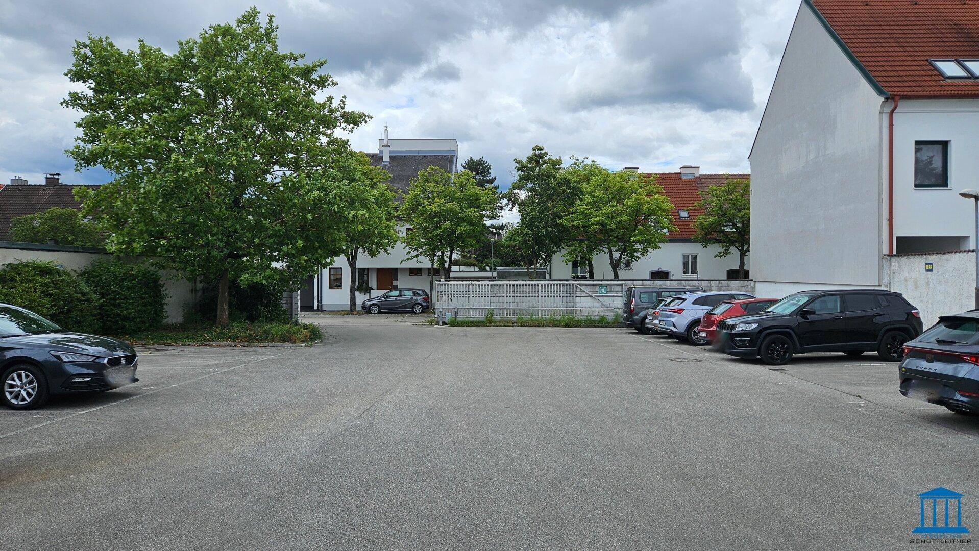 Außenparkplatz mit mehreren geparkten Autos und Bäumen im Hintergrund, unter bewölktem Himmel.