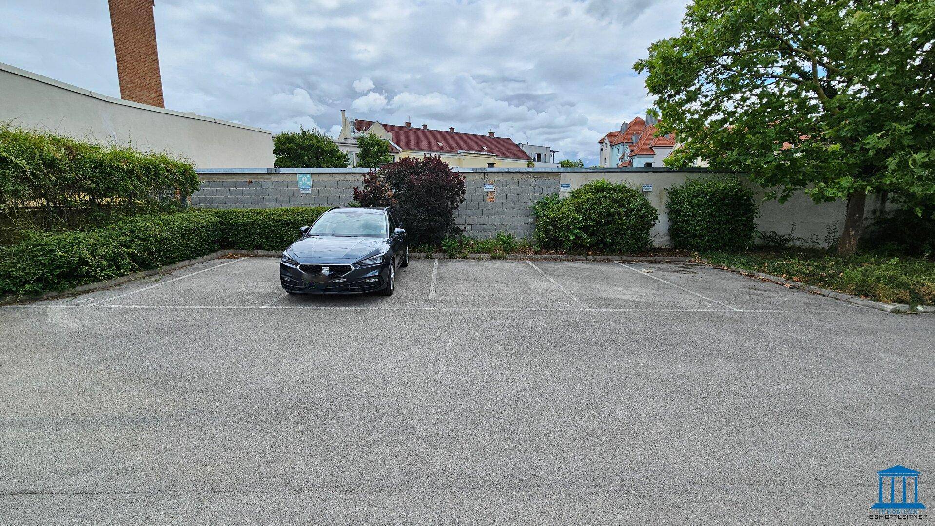 Außenparkplatz mit einem geparkten Auto, umgeben von Hecken und einer Mauer.