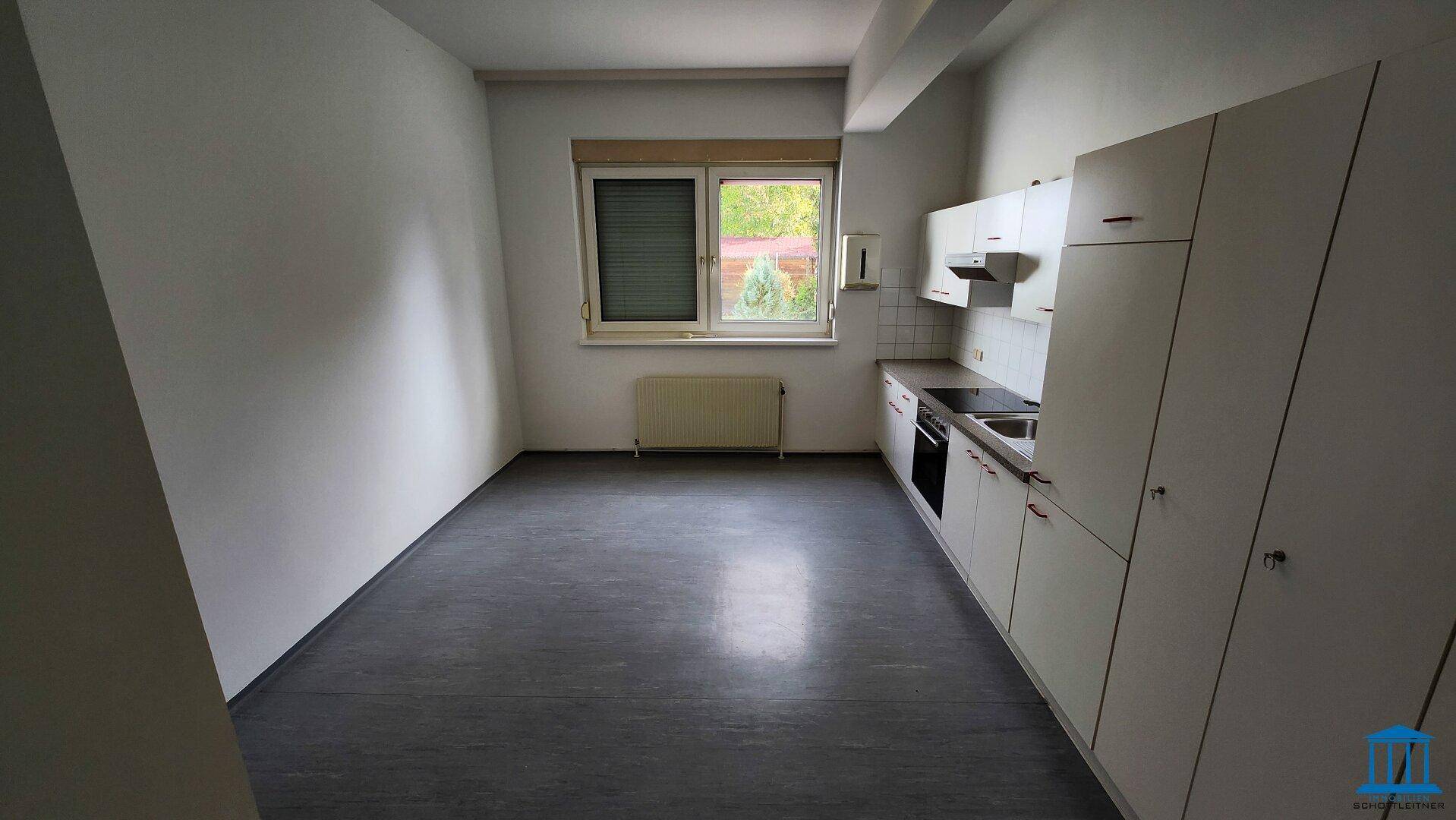 Kleine Küchenzeile mit weißen Schränken, Spüle, Herd und dunklem Vinylboden in einem hellen Raum.