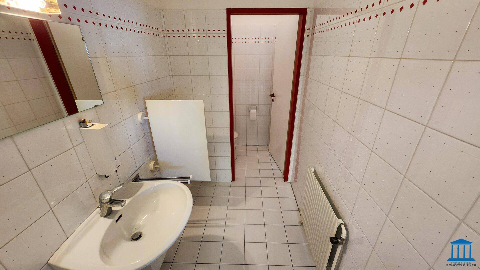 Toilette mit weißen Fliesen, Waschbecken und Spiegel, Zugang zu einem weiteren WC-Bereich.