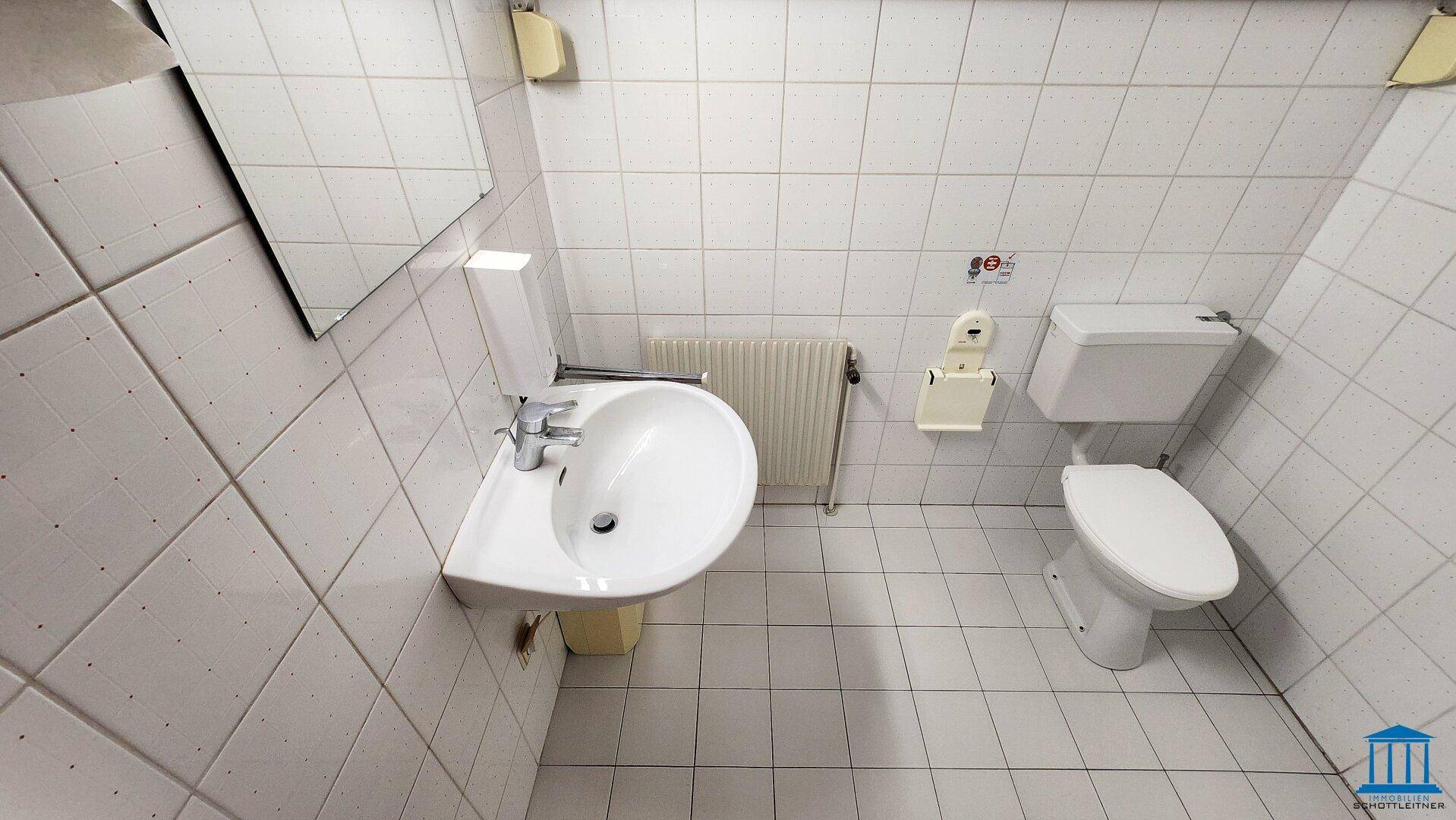 Einfache Toilette mit weißen Fliesen, Waschbecken und Heizkörper, sauber und funktional.