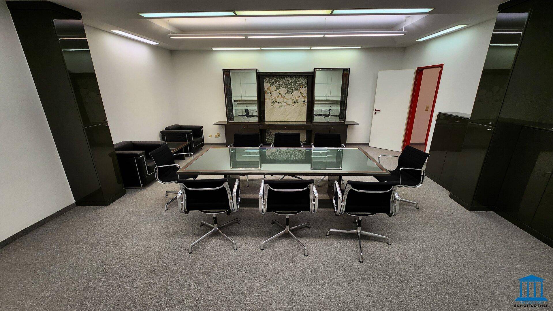 Geräumiger Konferenzraum mit elegantem Glastisch, schwarzen Ledersesseln und Teppichboden in dunklem Grau.