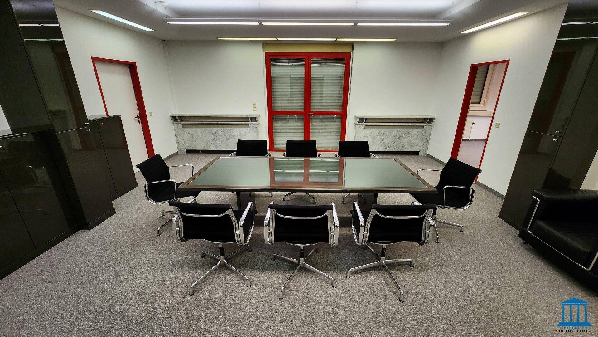 Konferenzraum mit großem Tisch, schwarzen Stühlen und Fenstern mit roten Rahmen, gut beleuchtet.