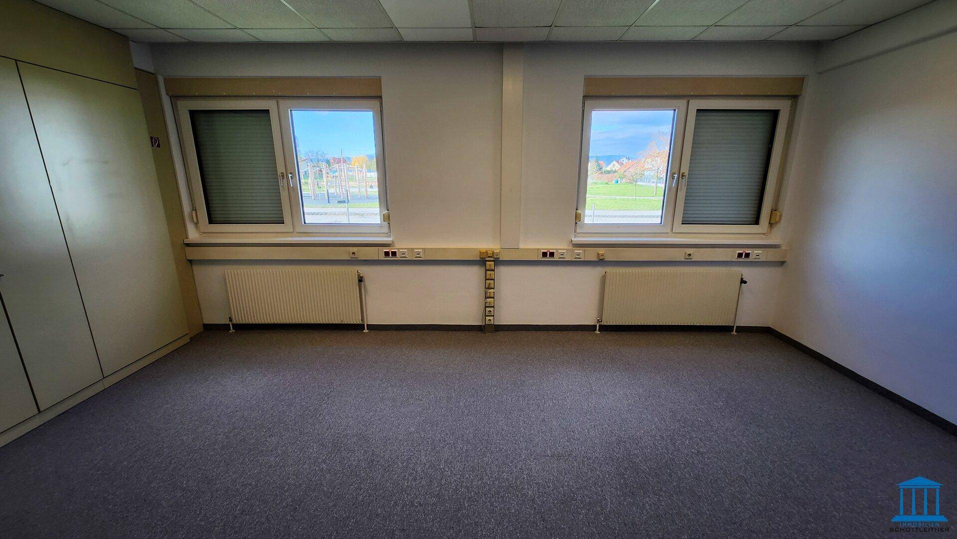 Helles Büro mit zwei Fenstern, Teppichboden und Heizkörpern unter den Fenstern, gut beleuchtet.