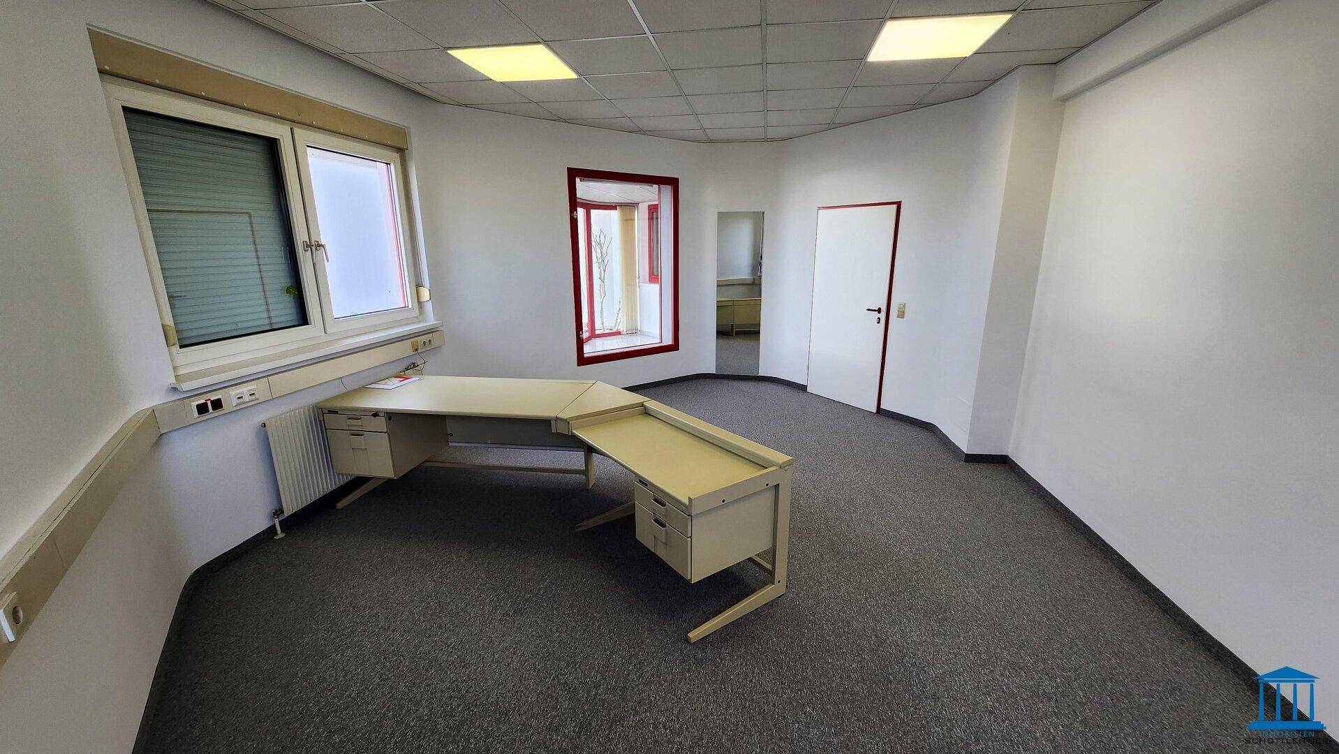 Büro mit Teppichboden, Eckschreibtisch und Blick auf einen weiteren Raum durch eine Tür.