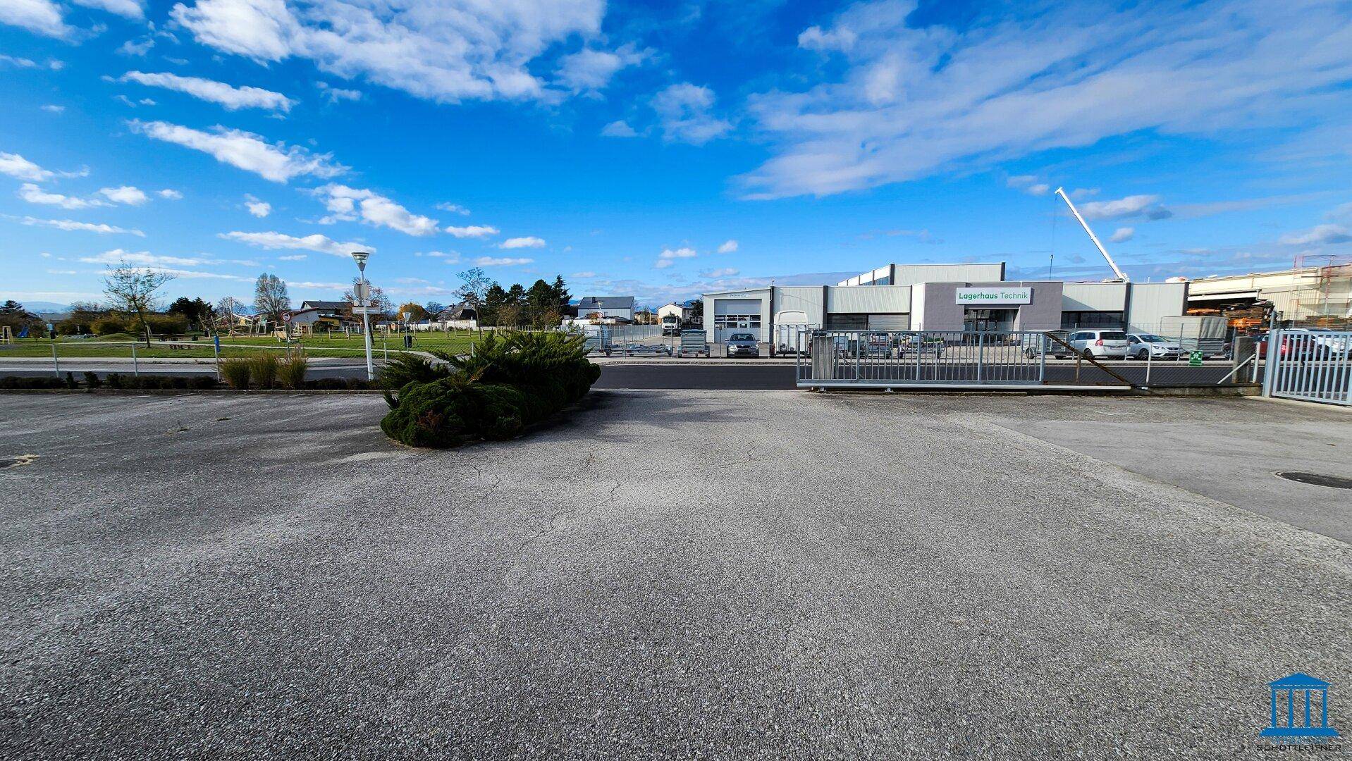 Weitläufiger Parkplatz mit Blick auf angrenzende Gewerbegebäude und blauen Himmel, sehr offen.