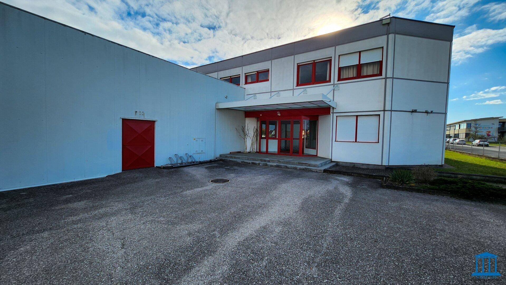 Frontansicht des Gewerbegebäudes mit Haupteingang und roter Tür, umgeben von einem Parkplatz.