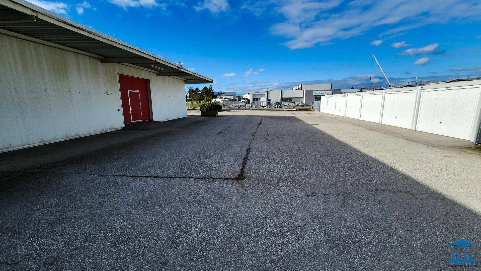 Asphaltierter Parkplatz neben dem Gewerbegebäude mit roten Toren und Lagerboxen im Hintergrund.