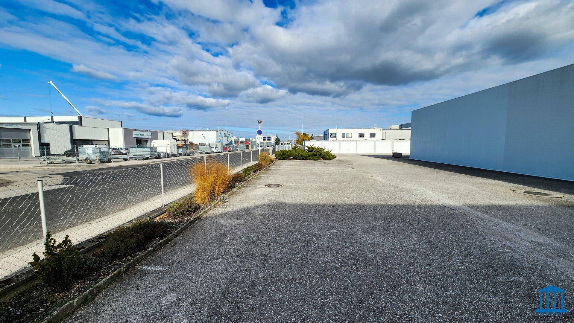 Asphaltierter Parkplatz mit Zaun und Blick auf weitere Gewerbegebäude in der Umgebung.