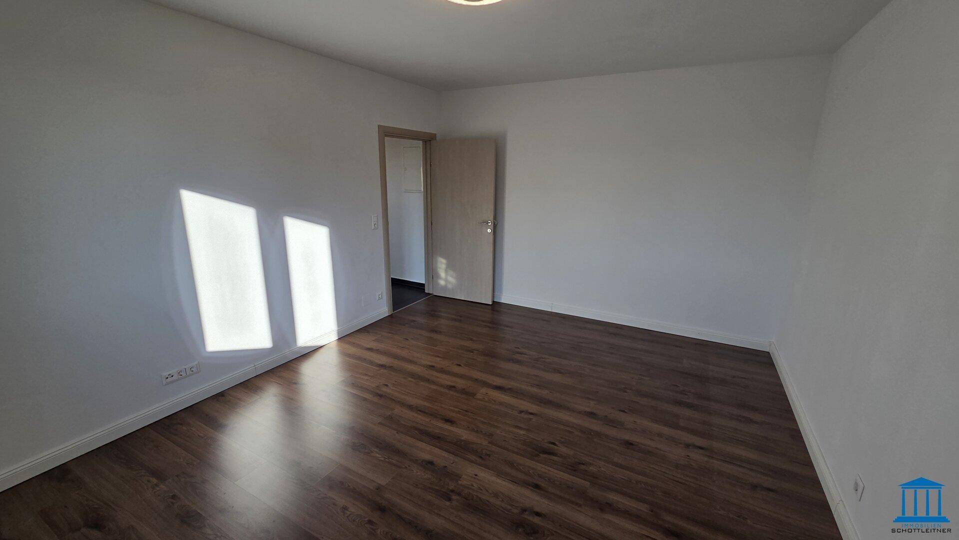 Helles Schlafzimmer mit dunklem Laminatboden und einer Tür, die zu einem weiteren Raum führt.